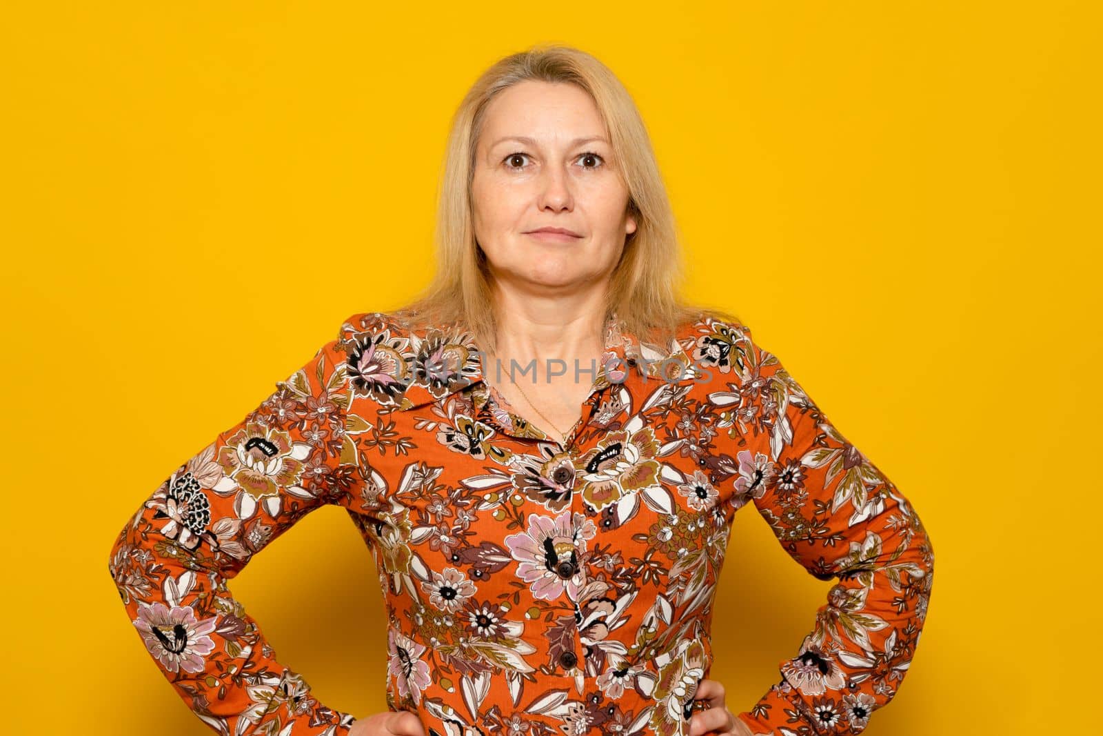 Smiling woman in patterned dress posing isolated on yellow wall background studio portrait. People emotions lifestyle concept. Mock up copy space. Standing with arms akimbo on the waist.