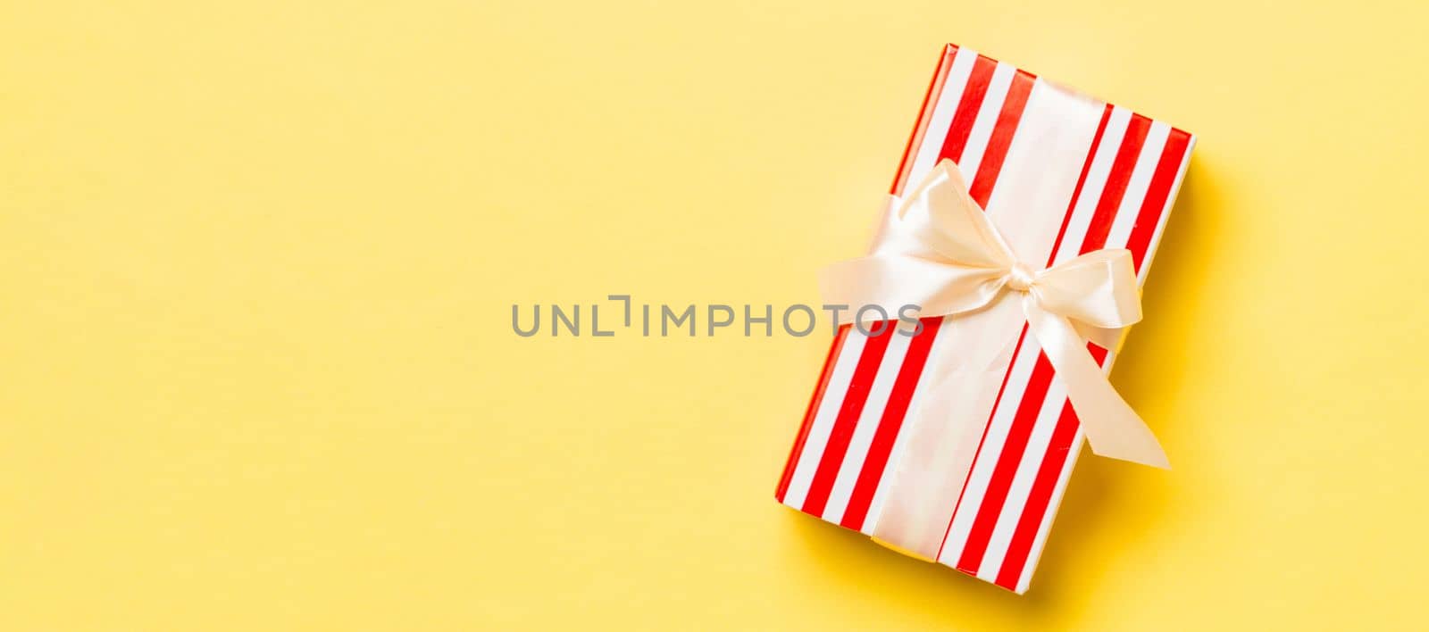 wrapped Christmas or other holiday handmade present in paper with yellow ribbon on yellow background. Present box, decoration of gift on colored table, top view with copy space.