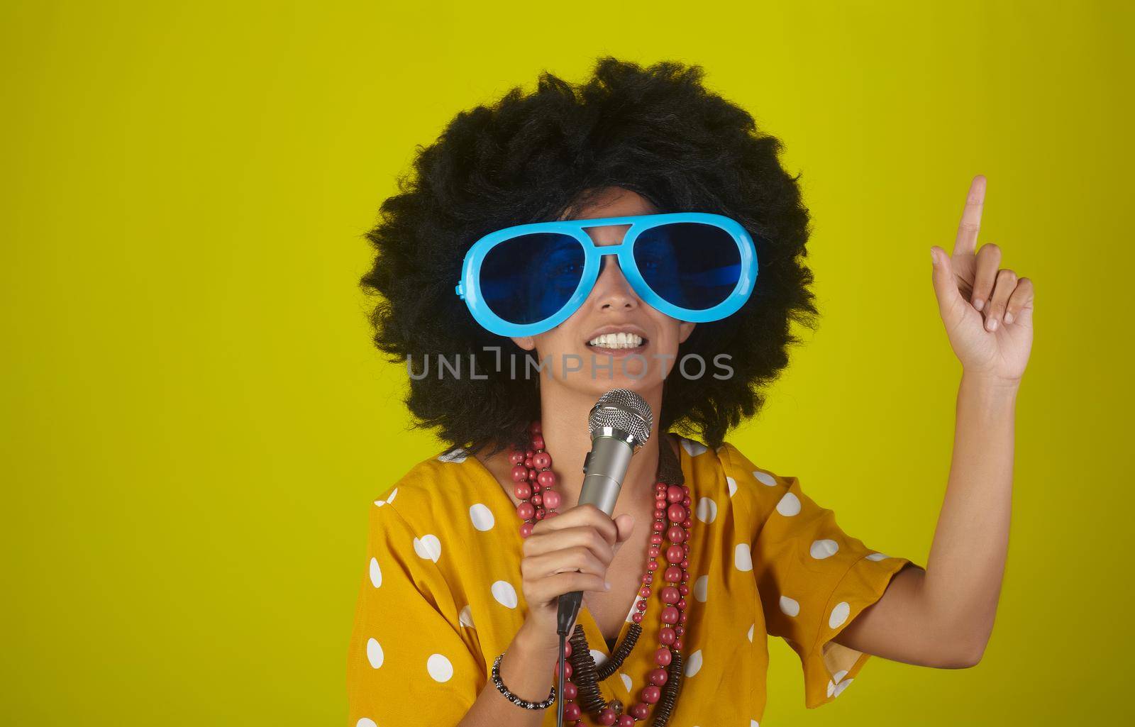 Young beautiful and smiling woman with the curly afro hairstyle and funny sunglasses singing using a microphone on yellow background