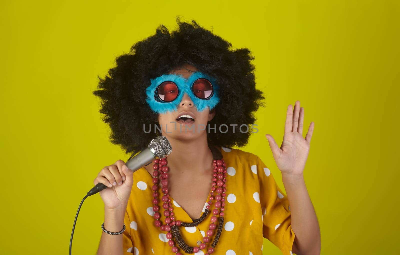 Young beautiful and smiling woman with the curly afro hairstyle and funny sunglasses singing using a microphone on yellow background
