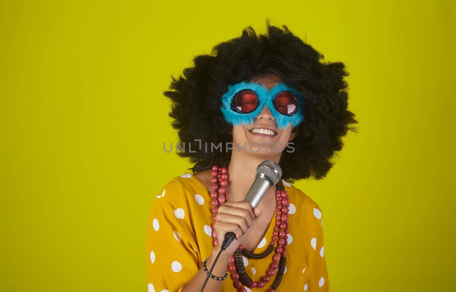 Young beautiful and smiling woman with the curly afro hairstyle and funny sunglasses singing using a microphone on yellow background