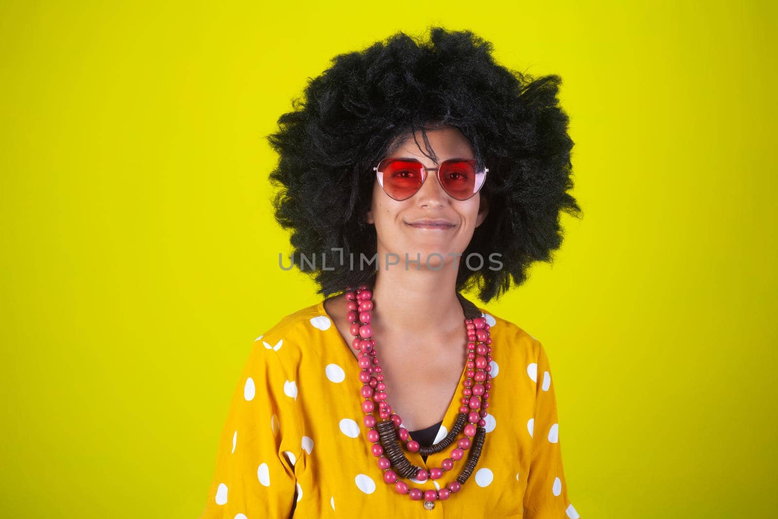 Portrait of a smiling woman with the afro curly hairstyle wearing the heart shaped sunglasses on yellow background