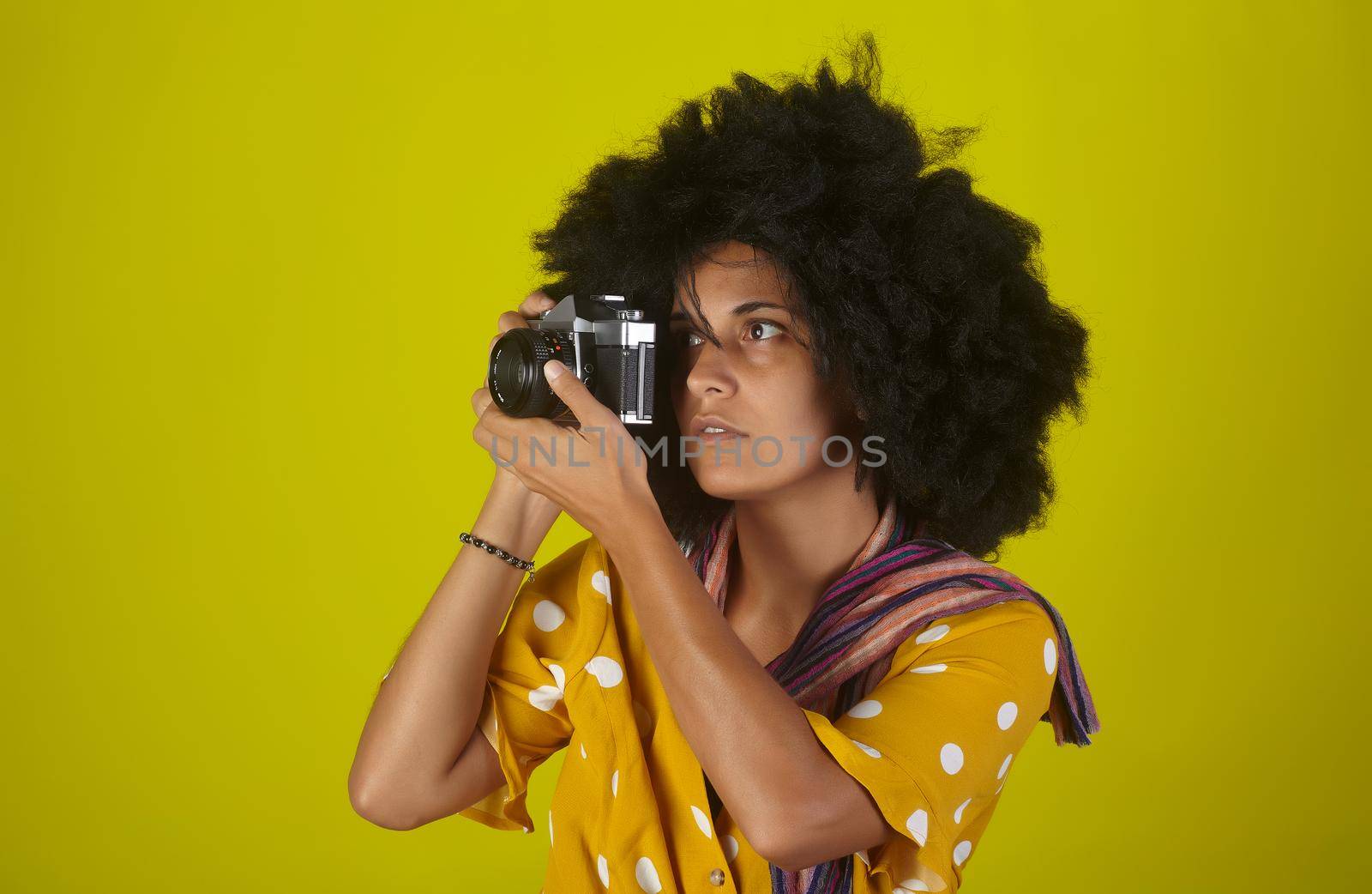 A beautiful woman with curly afro hairstyle on yellow background while taking pictures with a retro films camera