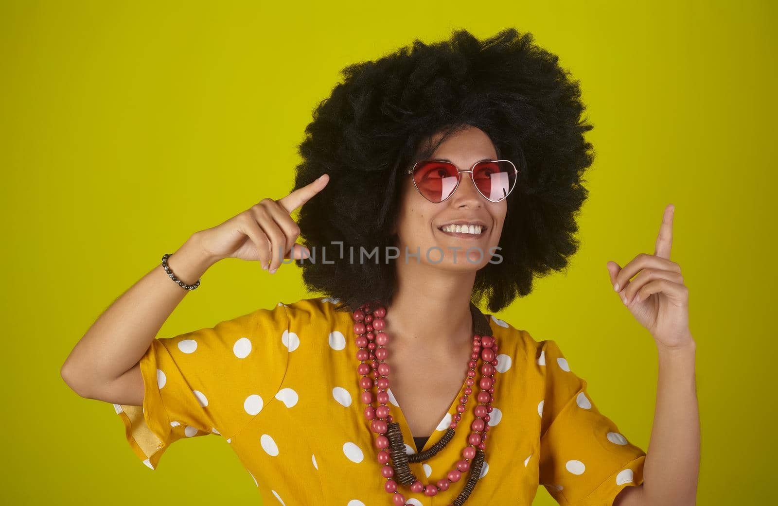 A beautiful and smiling woman with the curly afro hairstyle wearing heart shaped sunglasses looking and indicating direction up to the right with fingers on yellow background