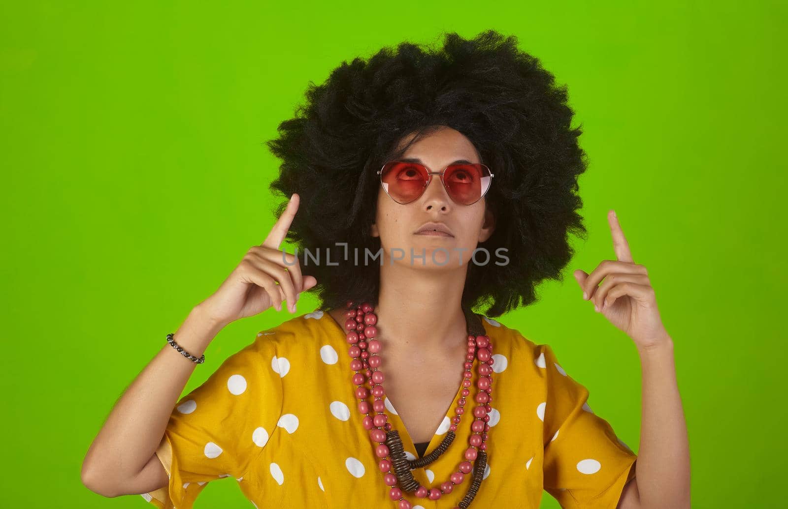 A beautiful woman with the curly afro hairstyle wearing heart shaped sunglasses looking and indicating direction up to the right with fingers on green background