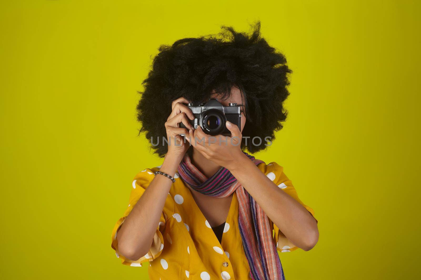 A beautiful woman with curly afro hairstyle on yellow background while taking pictures with a retro camera