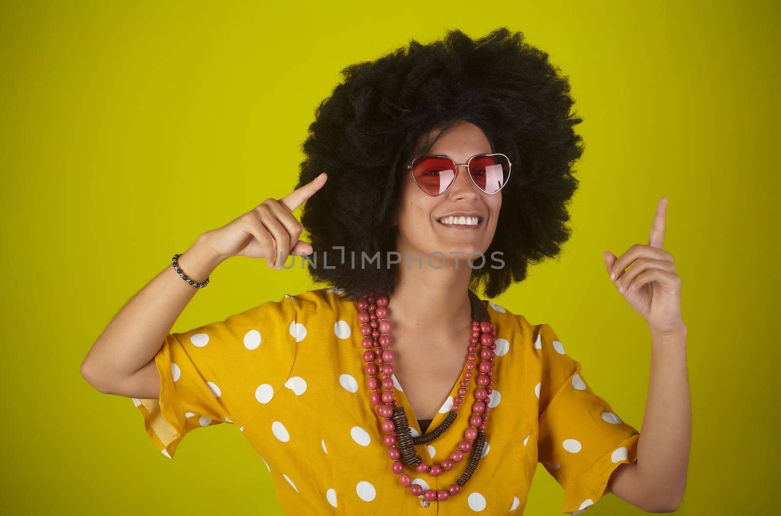 A beautiful and smiling woman with the curly afro hairstyle wearing heart shaped sunglasses looking and indicating direction up to the right with fingers on yellow background