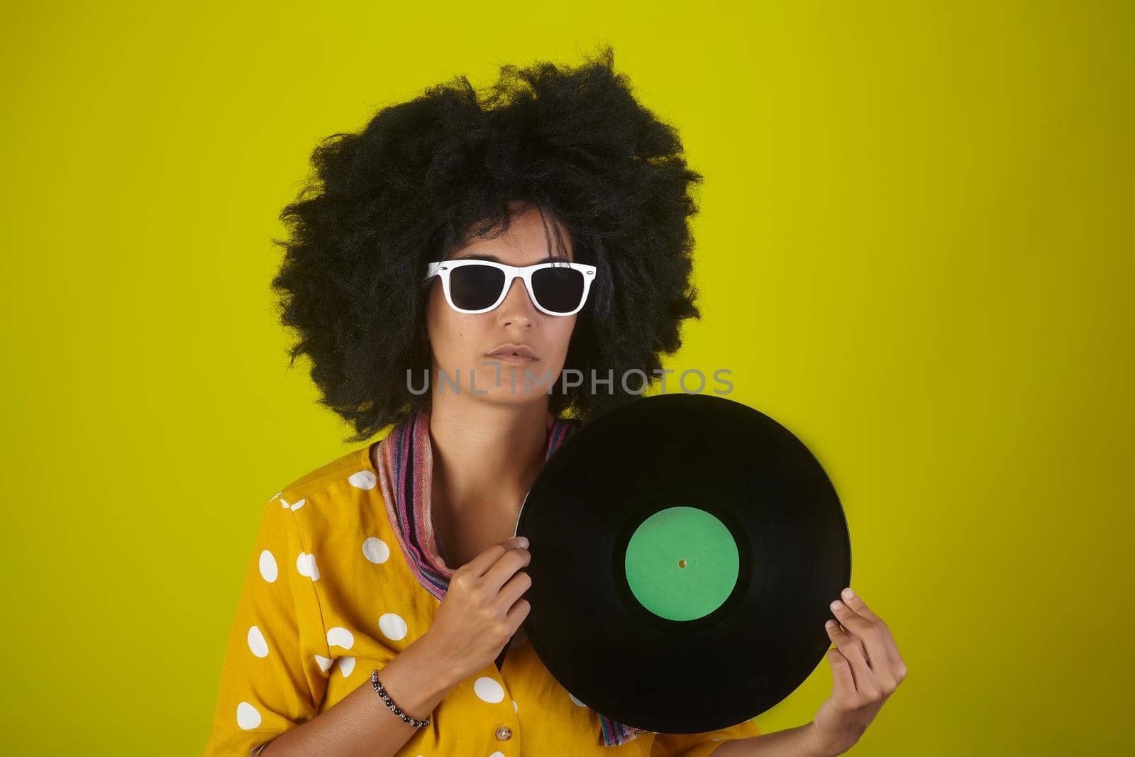 A woman with sunglasses and afro hairstyle holding a vinyl disk on a yellow background