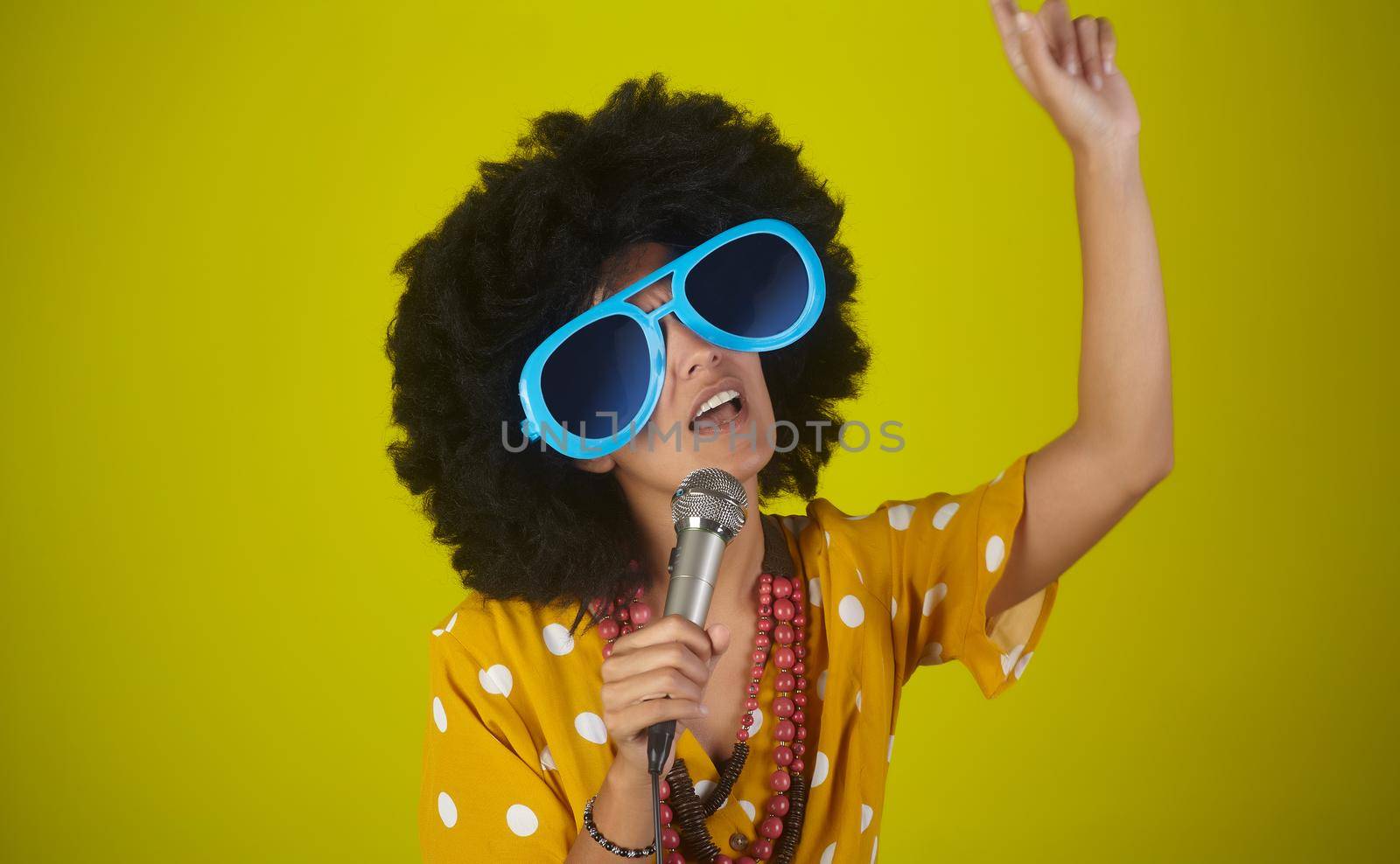 Young beautiful and smiling woman with the curly afro hairstyle and funny sunglasses singing using a microphone on yellow background