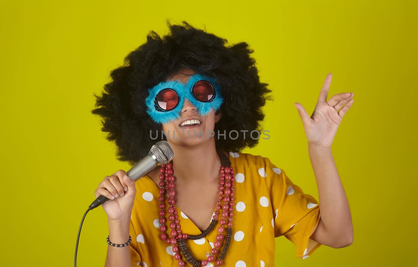 Young beautiful and smiling woman with the curly afro hairstyle and funny sunglasses singing using a microphone on yellow background