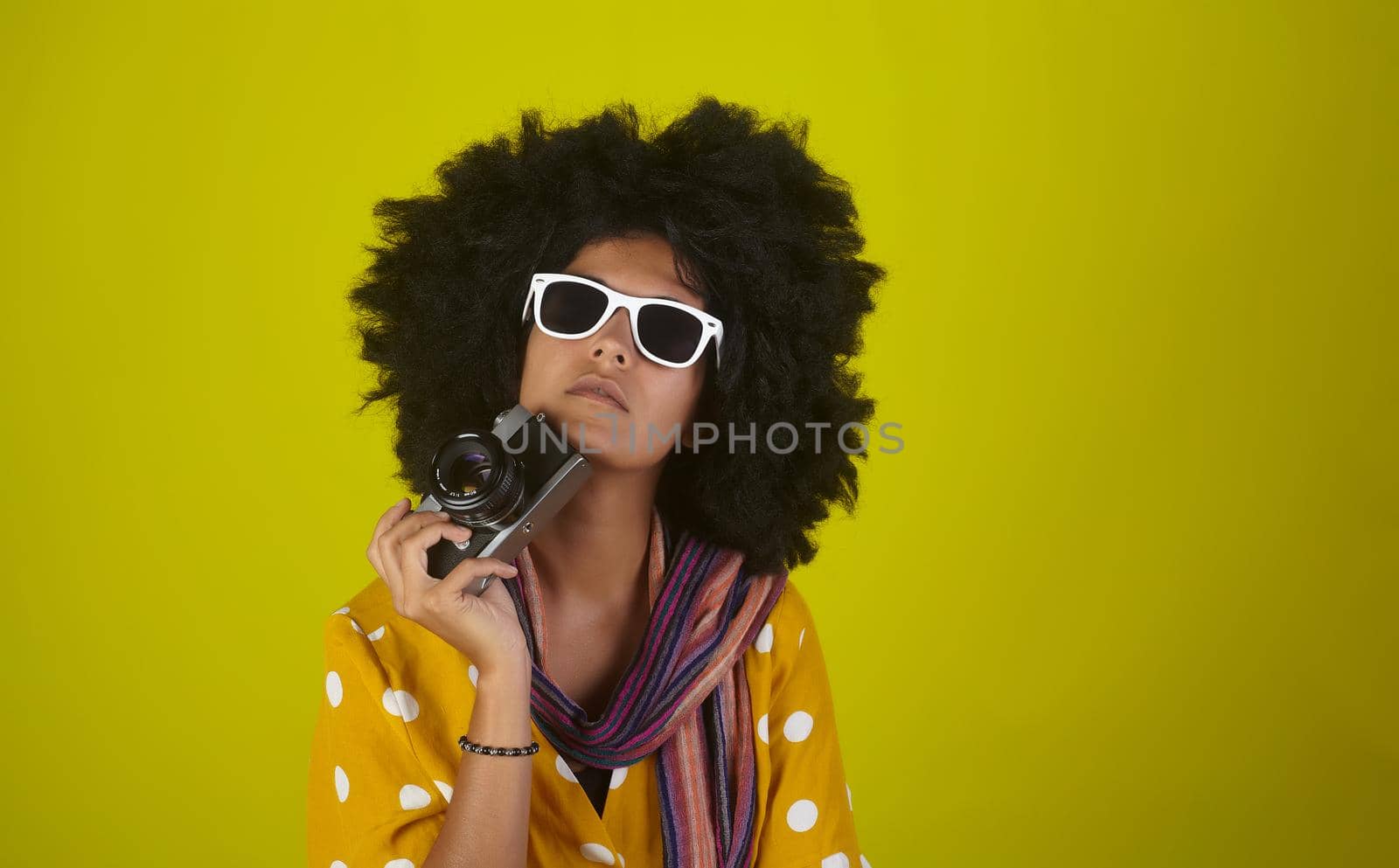 A beautiful woman with curly afro hairstyle holkding a retro film camera on yellow background