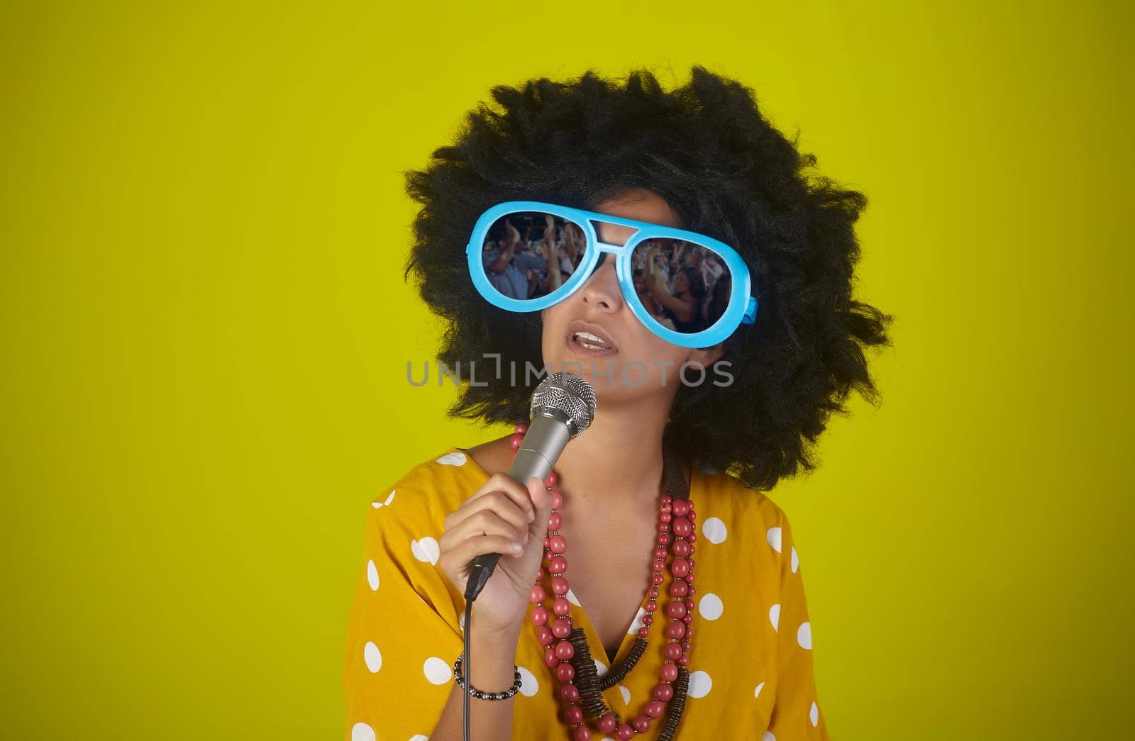 A beautiful woman with the curly afro hairstyle and funny sunglasses singing using a microphone on yellow background