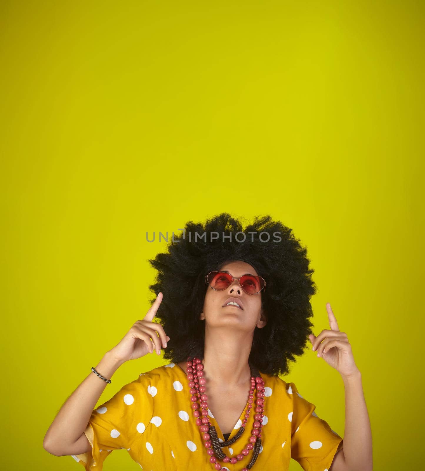 Beautiful woman with the curly afro hairstyle wearing heart shaped sunglasses looking and indicating direction up to the right with fingers on yellow background by bepsimage