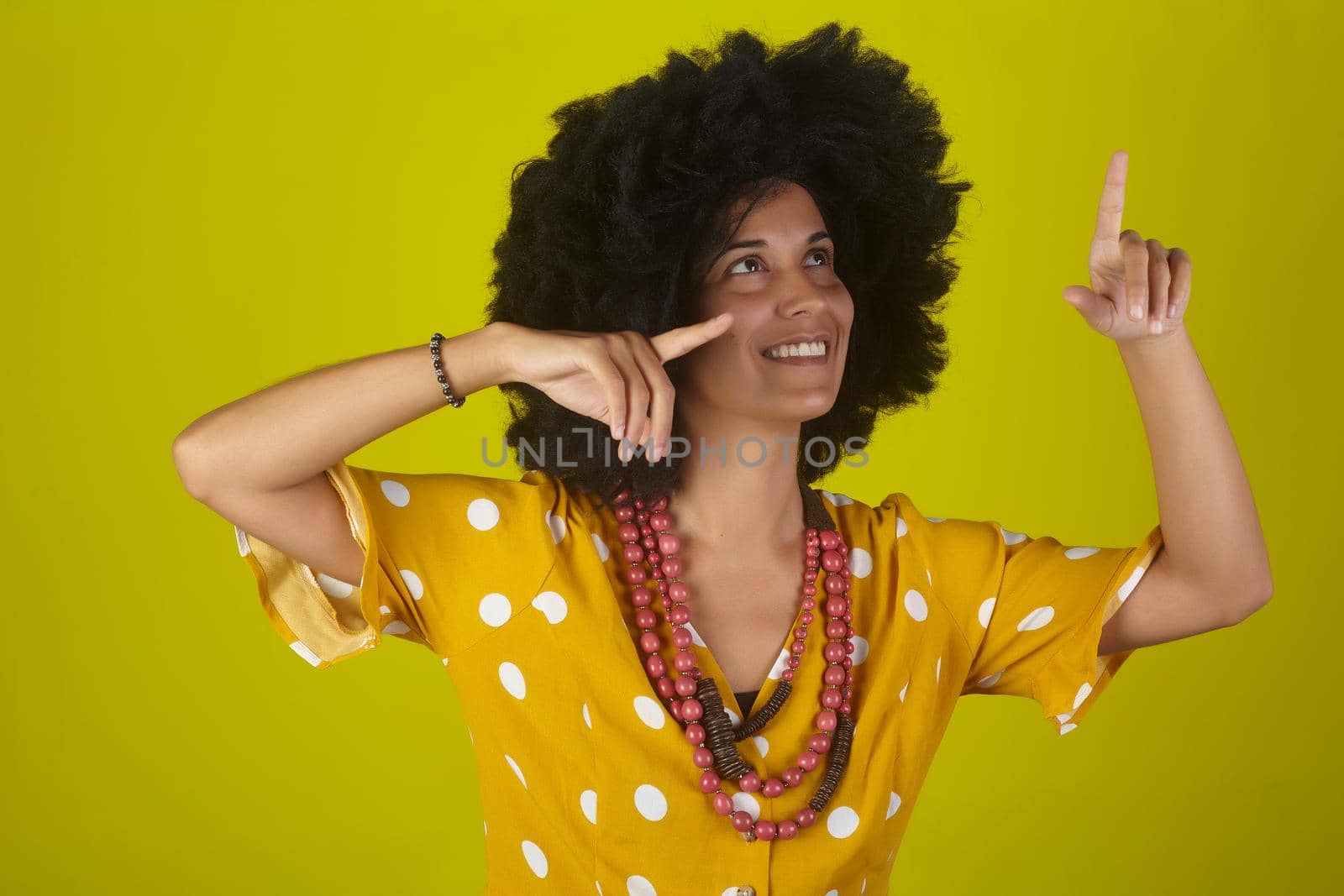 A beautiful and smiling woman with the curly afro hairstyle looking and indicating direction up to the right with fingers on yellow