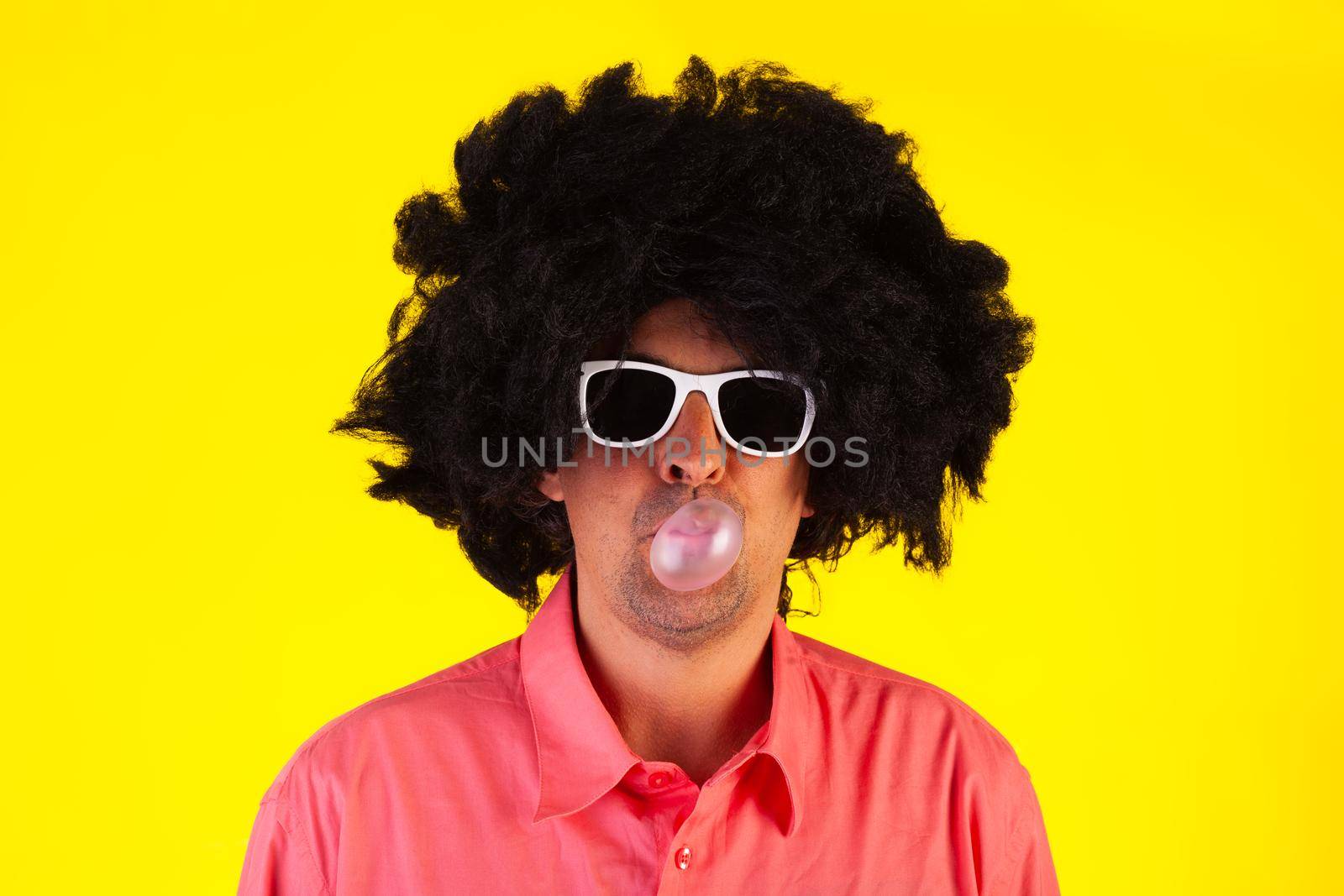 Curly man wearing sunglasses blowing bubble with chewing gum on yellow background