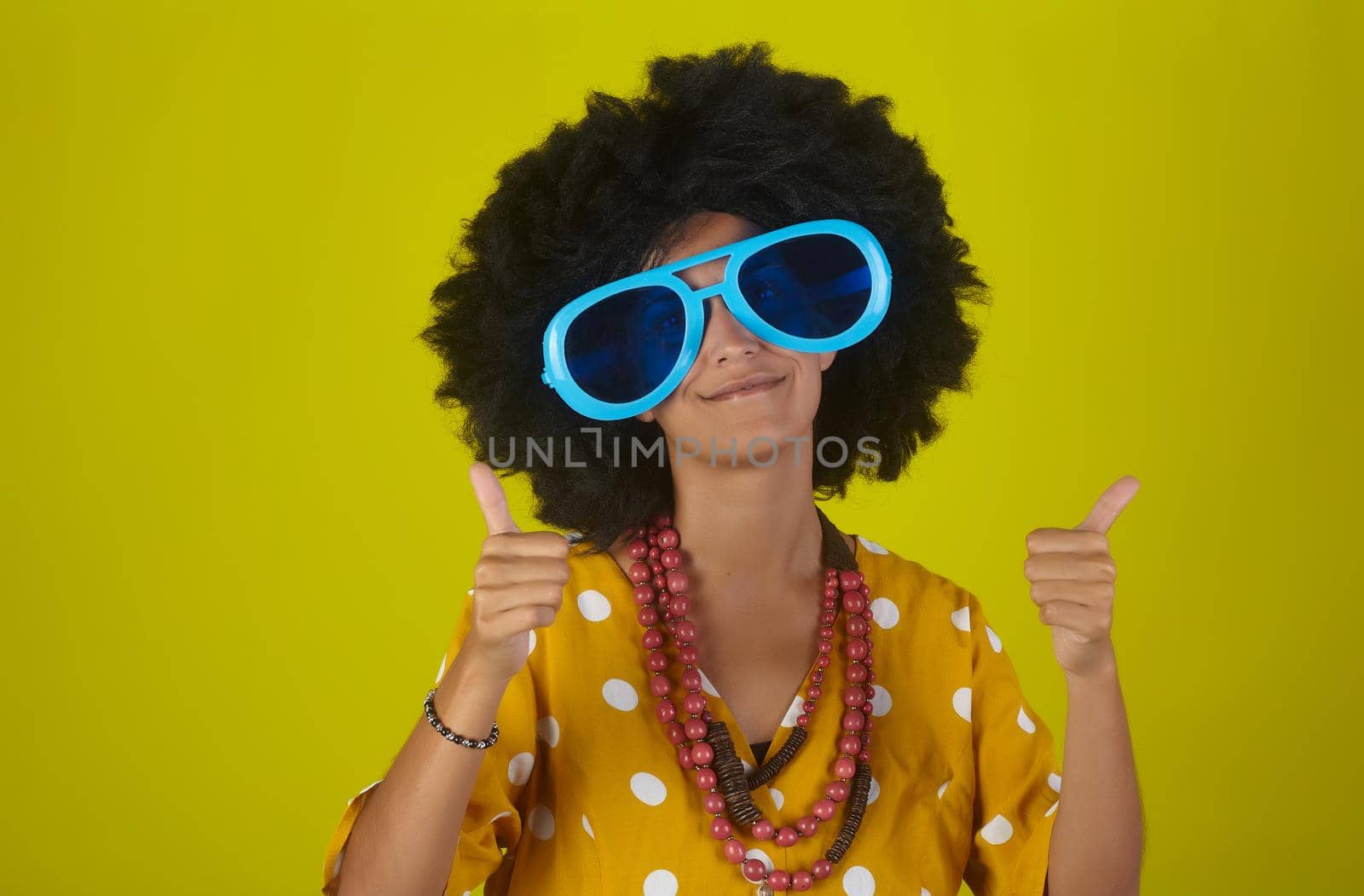 Beautiful and smiling woman with curly afro hairstyle and funny sunglasses showing thumbs up on yellow background by bepsimage