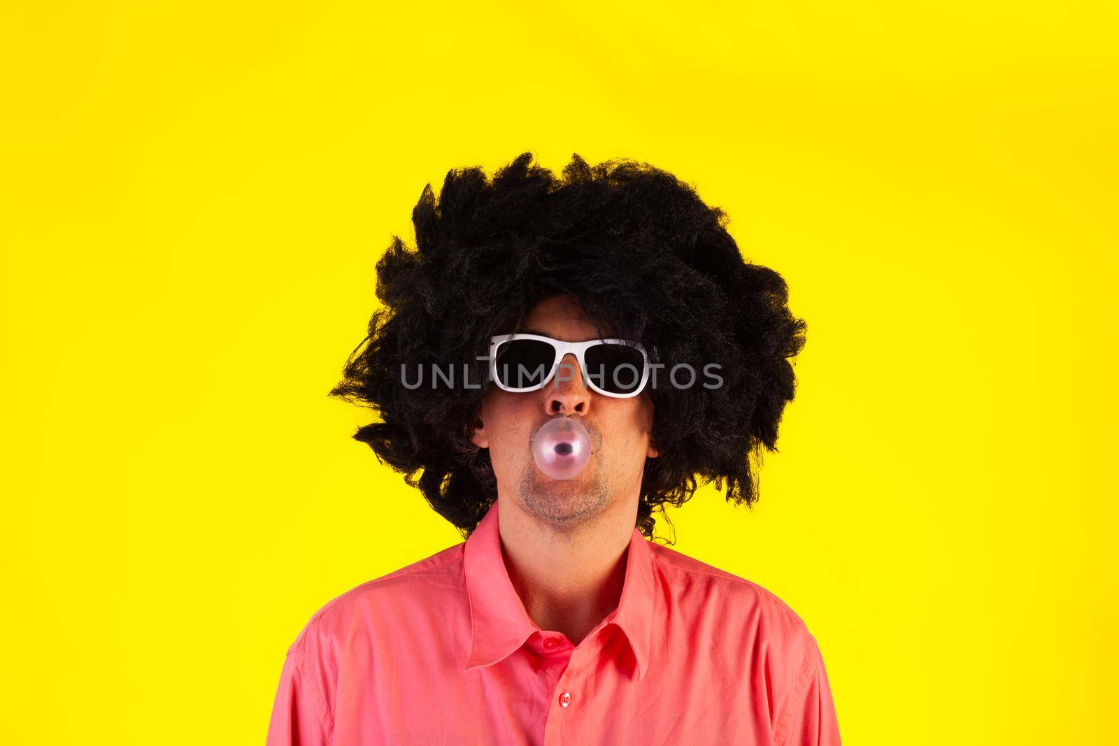 Curly man wearing sunglasses blowing bubble with chewing gum on yellow background