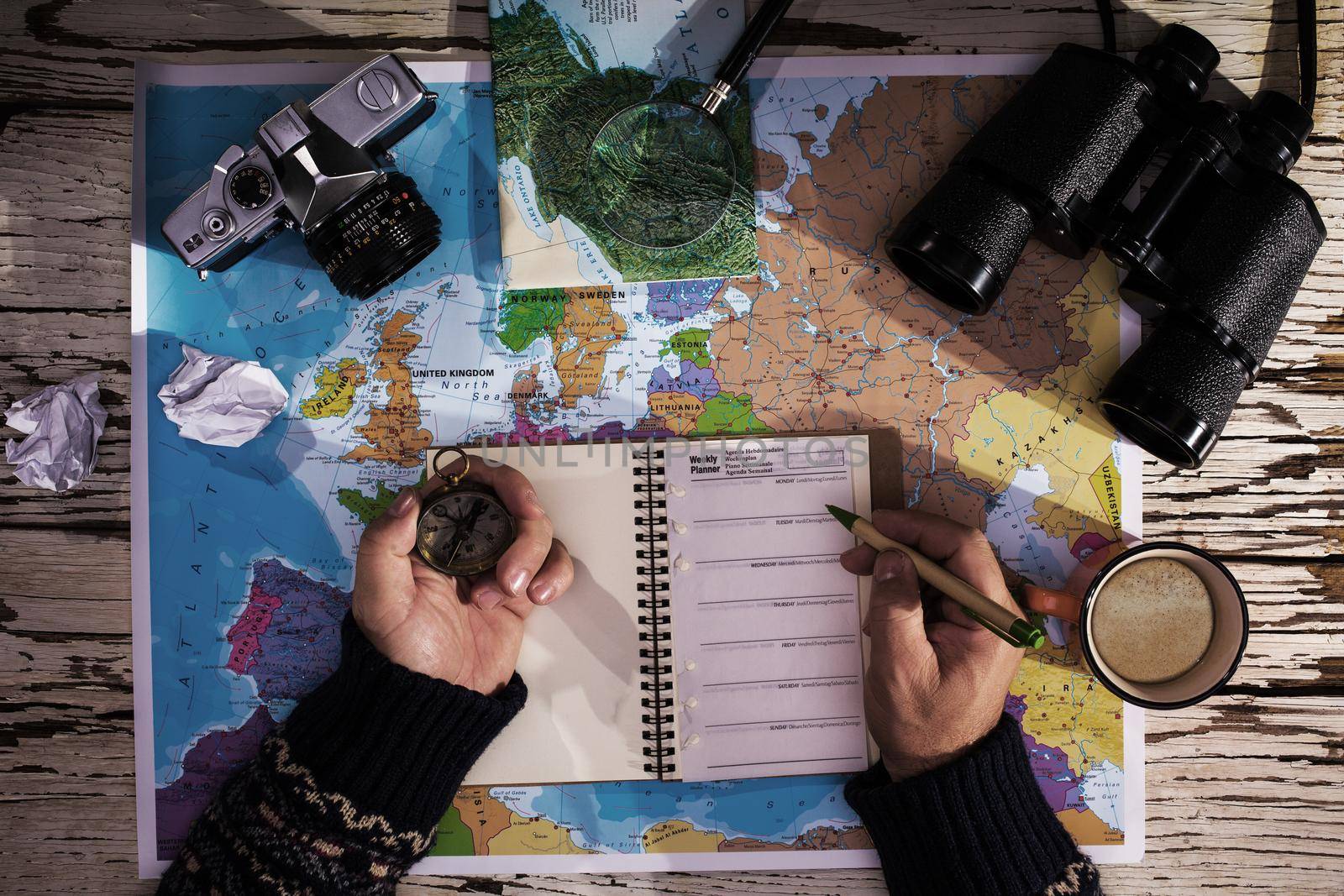 Flat lay of the travel planning concept. Top view of man hands wriìting on a diary, on the table binoculars, compass, retro photo camera, coffee, and europe map on a white wooden table