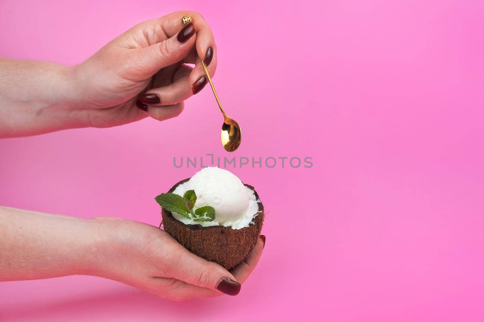 Vanilla ice cream balls in a fresh coconut half, decorated with mint leaves in the hand with a spoon on a pink background.