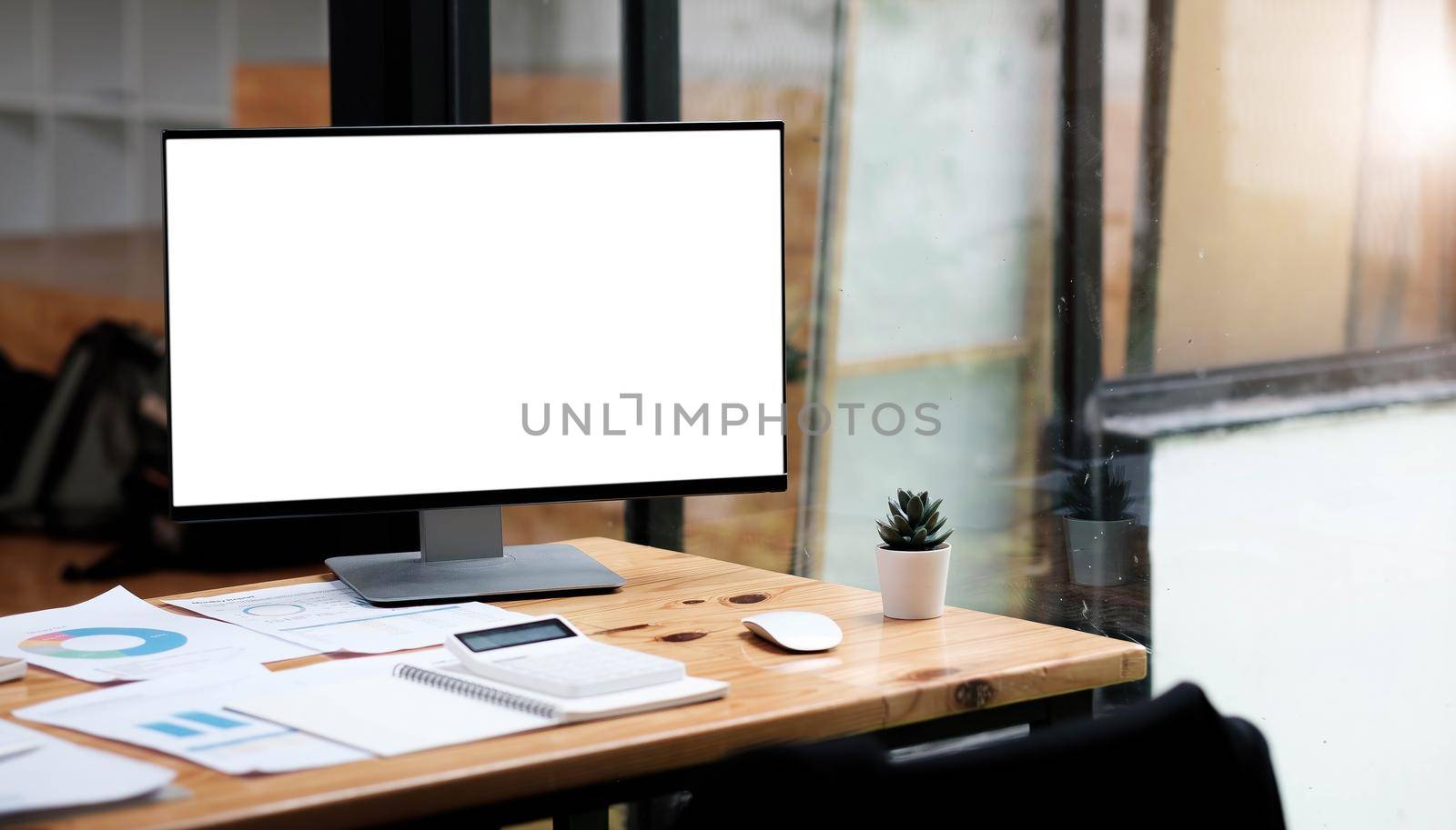 Computer laptop with white blank screen putting on wooden working desk that surrounded by coffee cup, stack of books, potted plant, pencils over comfortable living room windows as background..