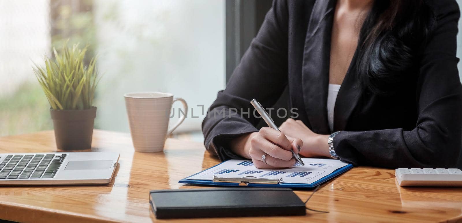 Close up of business woman working at office desk. Right hand writing in notebook. Laptop, eyeglasses calculator and business summary charts on background. by wichayada