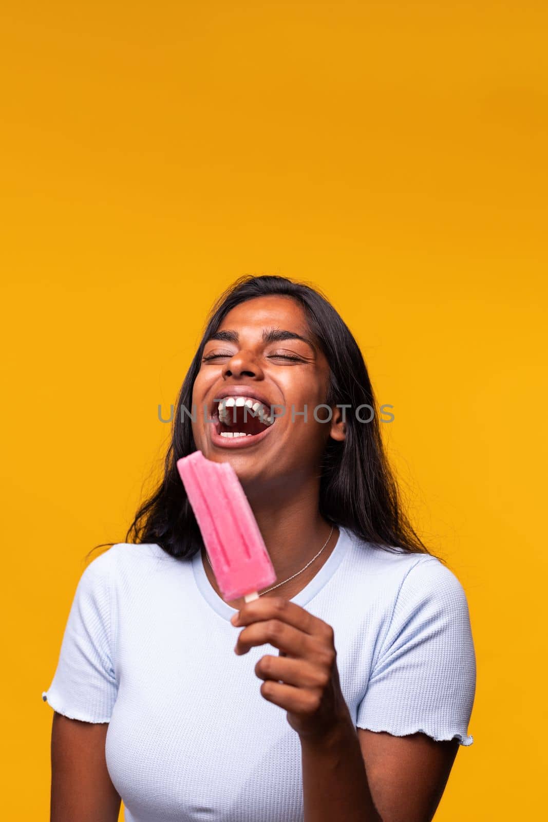 Vertical portrait of young Indian woman laughing eating pink popsicle on yellow background. Asian woman eating ice cream. Studio shot.
