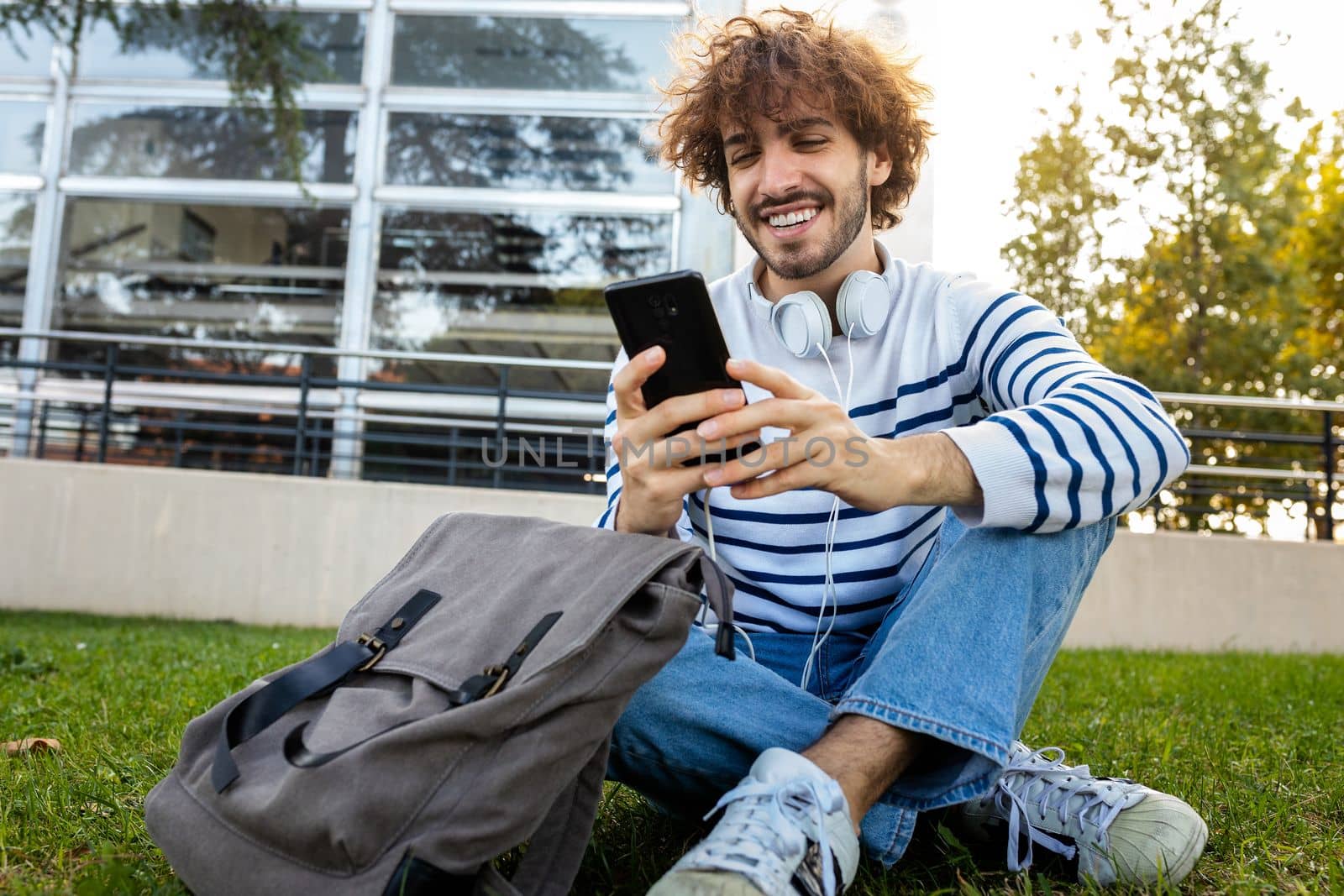 Happy young male university student sitting on grass in campus using mobile phone. Education concept.