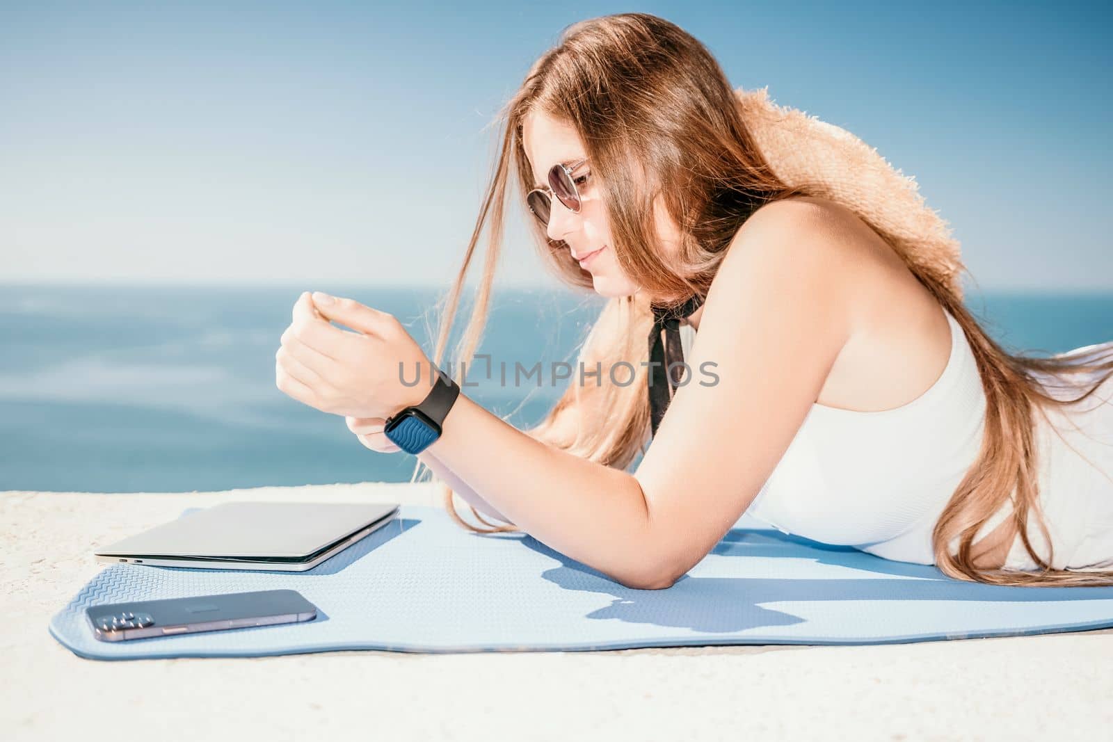 Successful business woman in yellow hat working on laptop by the sea. Pretty lady typing on computer at summer day outdoors. Freelance, travel and holidays concept.