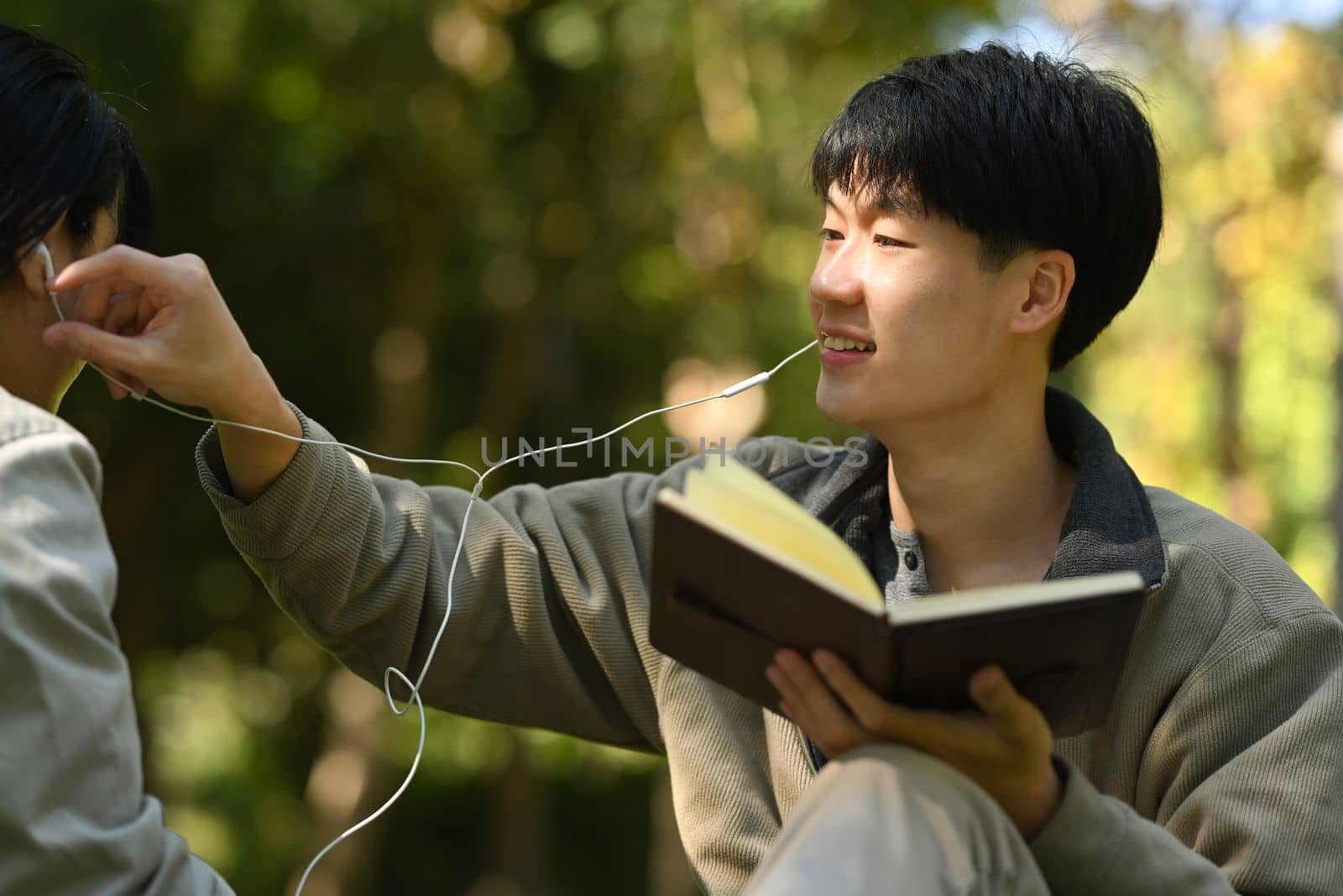 Image of young couple reading a book and listening to music in earphones while relaxing at public park together.