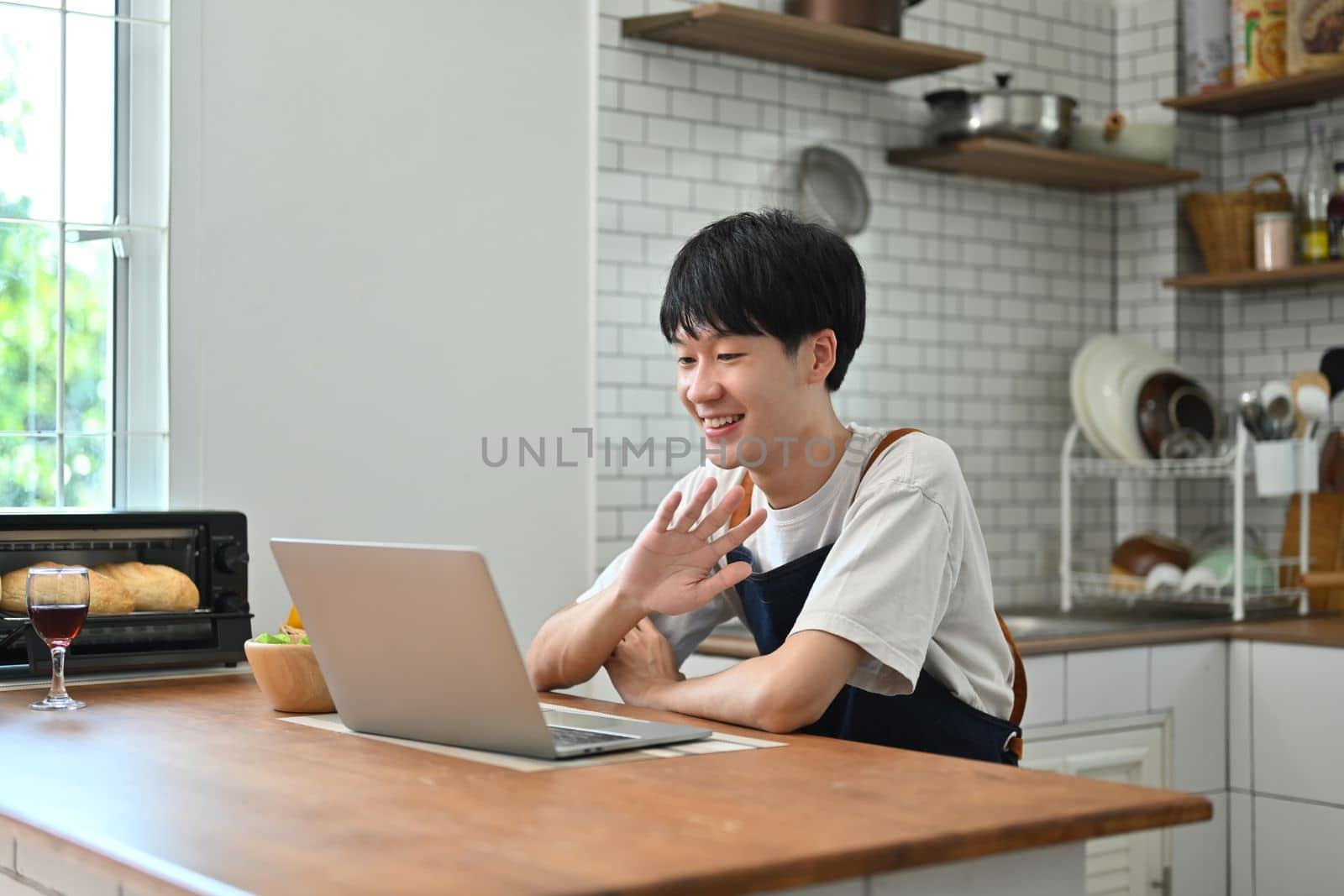 Cheerful young man freelancer checking email in morning, working online on laptop while sitting at wooden table in kitchen.