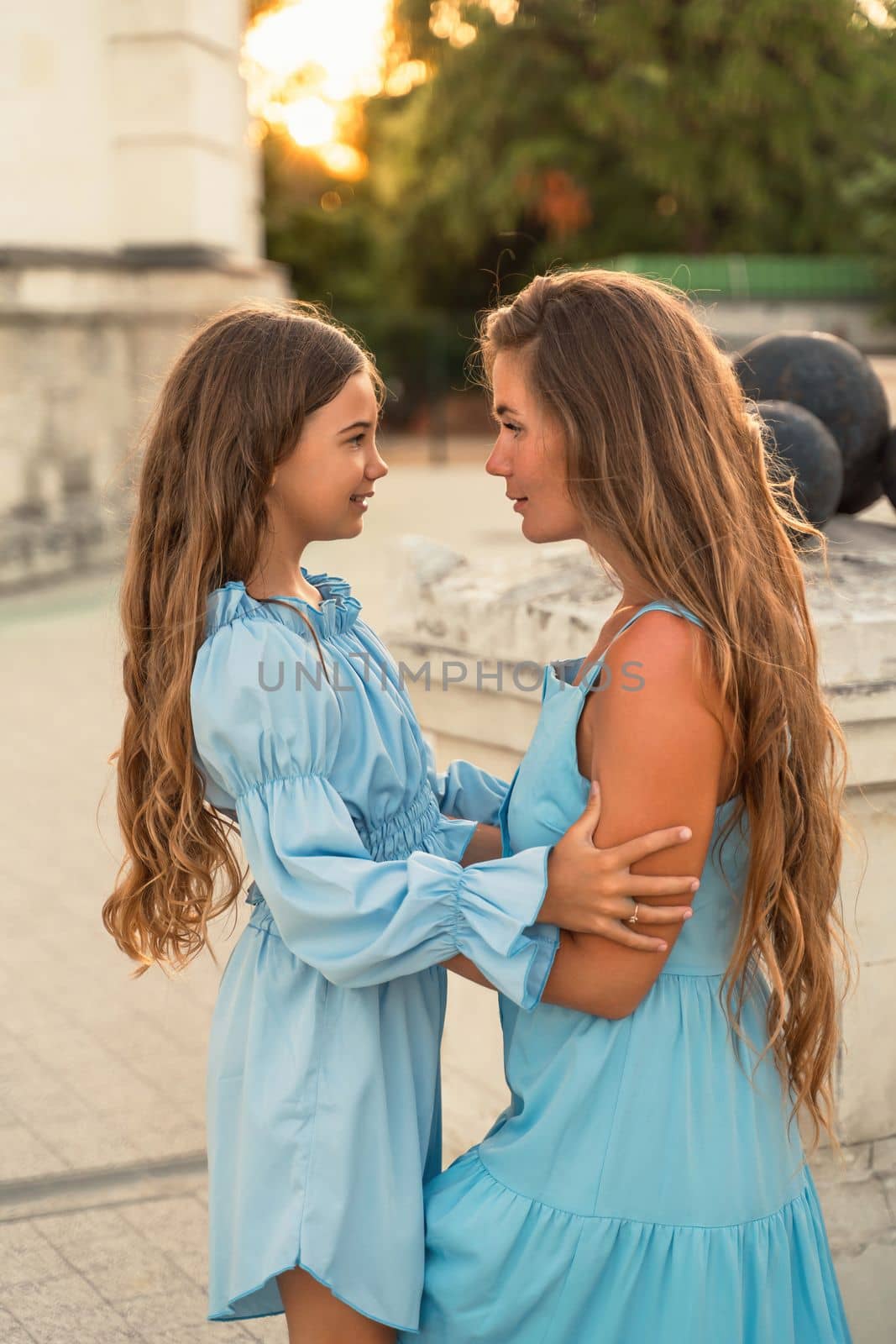 Portrait of a mother and daughter in blue dresses with flowing long hair against the backdrop of a sunset and a white building. They look at each other. Family stories on the weekend.