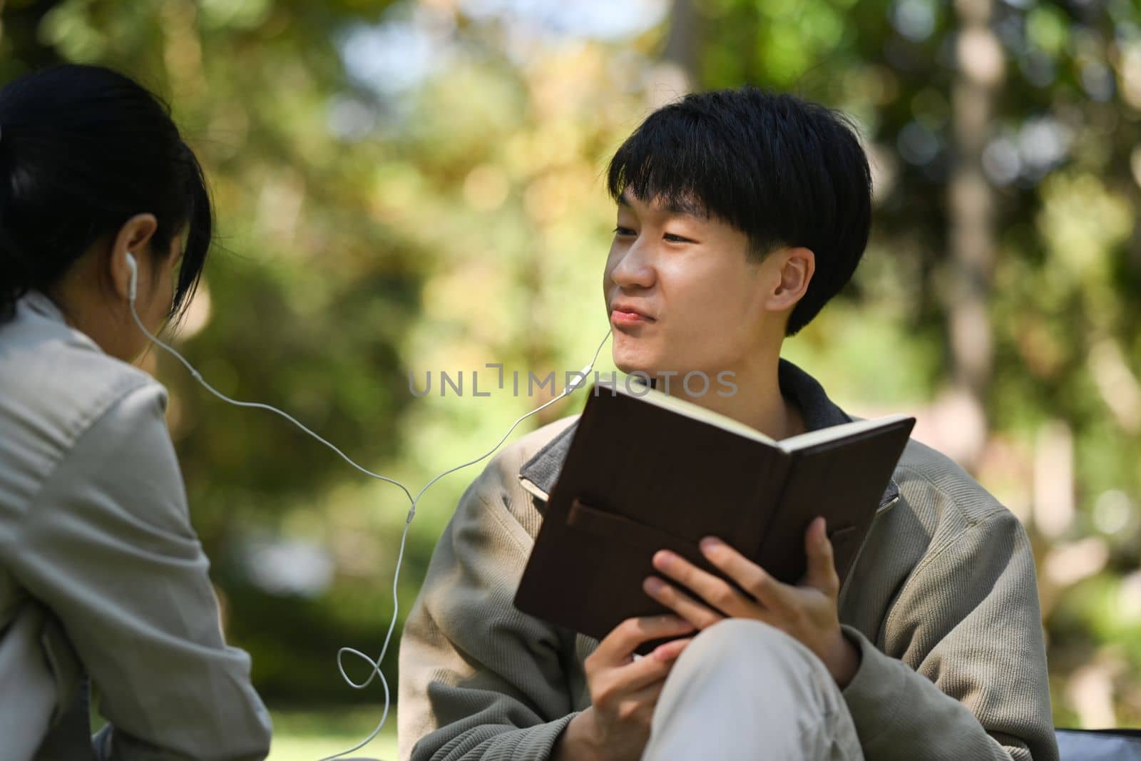 Image of young couple reading a book and listening to music in earphones while relaxing at public park together by prathanchorruangsak