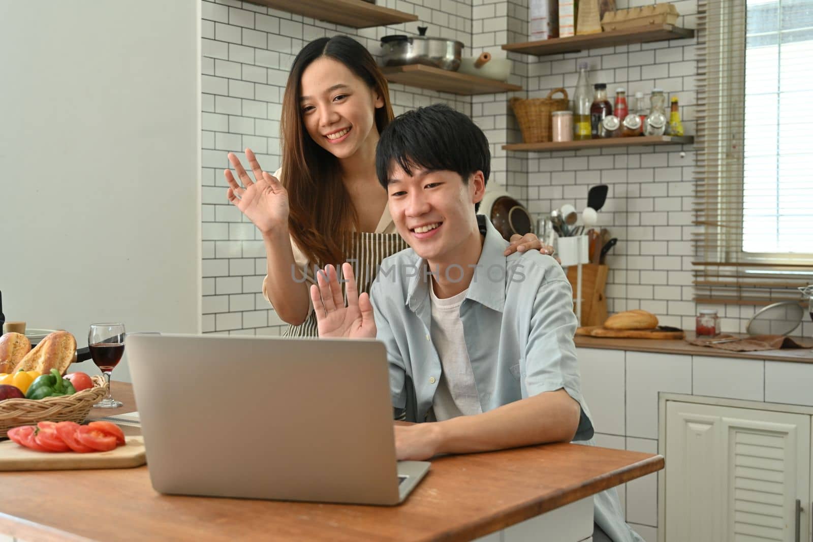 Image of adorable asian couple making video call with friends or family via laptop computer while sitting in cozy kitchen by prathanchorruangsak