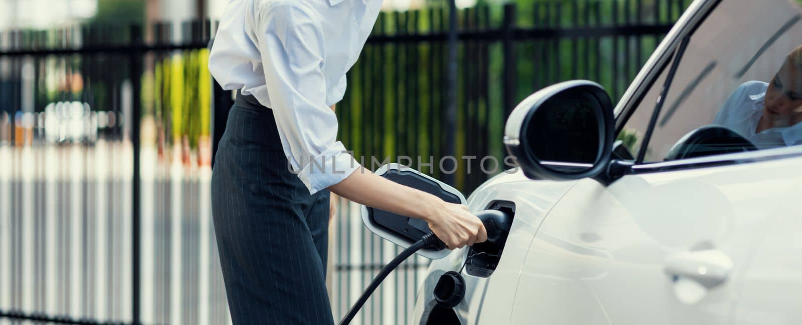 Closeup progressive suit-clad businesswoman with her electric vehicle recharge her car on public charging station in modern city with power cable plug and renewable energy-powered electric vehicle.