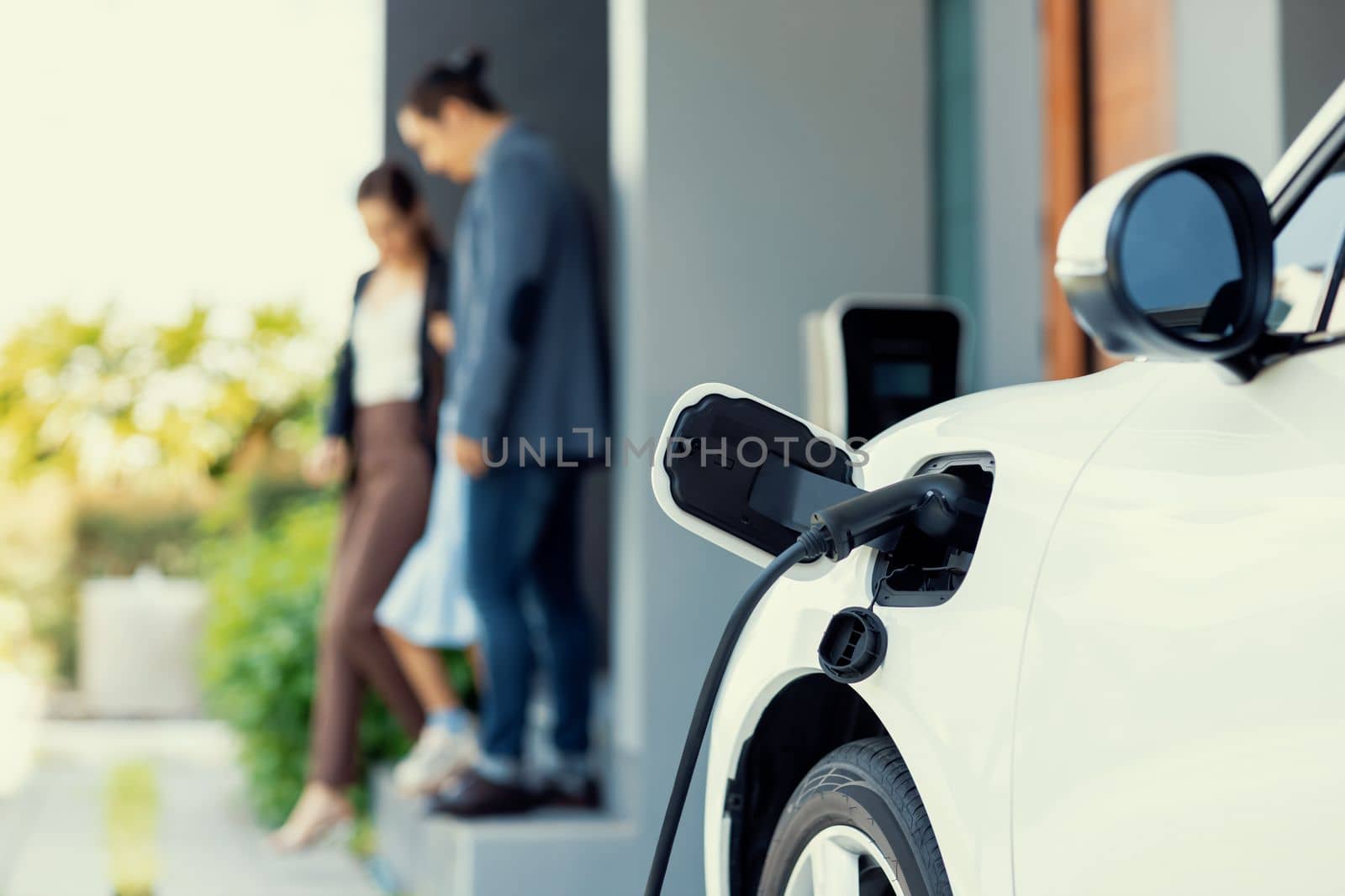 Focus closeup electric vehicle recharging battery from home electric charging station with blurred family in background. Renewable clean energy car for progressive eco awareness lifestyle concept.
