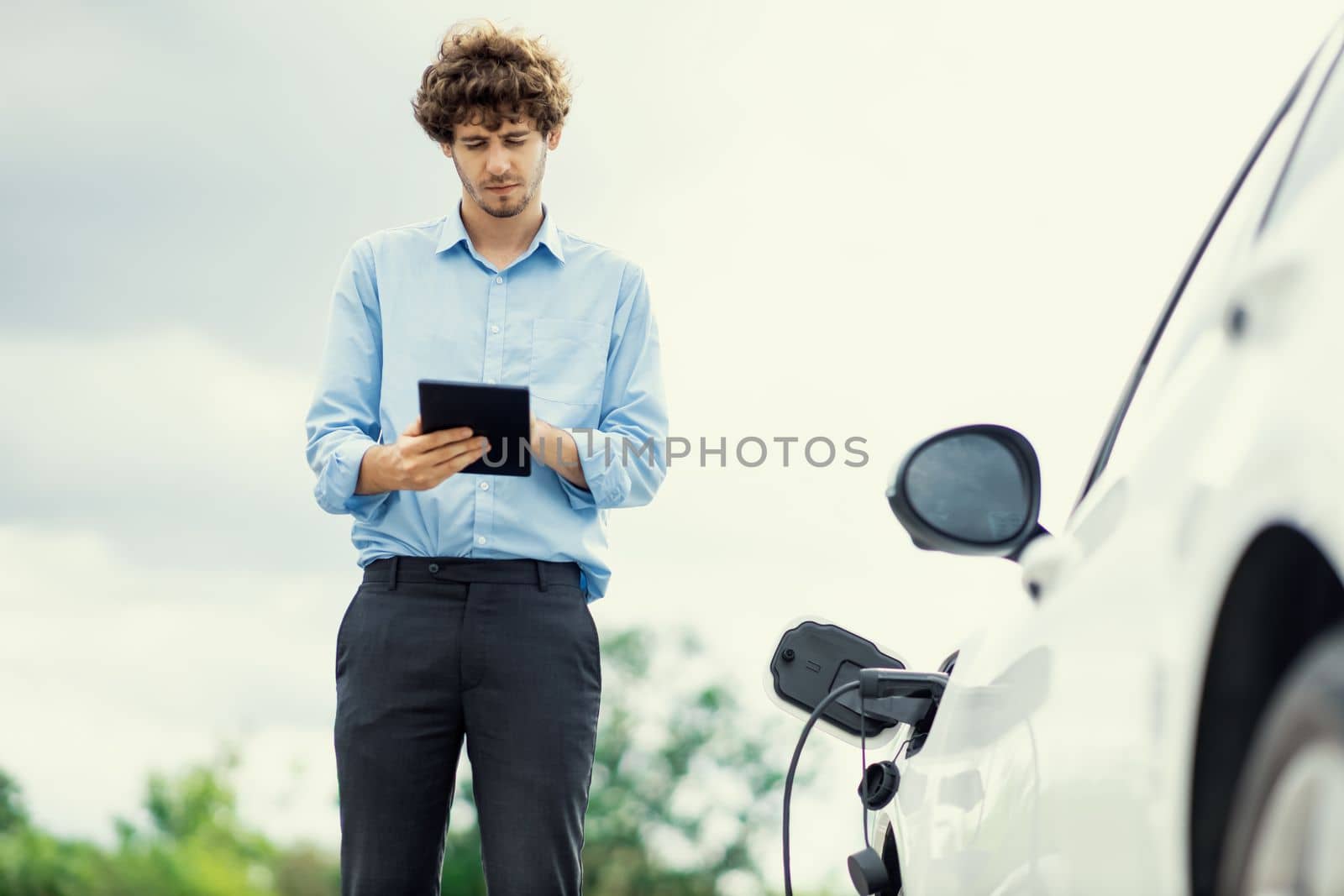 Closeup progressive suit-clad businessman with his electric vehicle recharge his car on public charging station in modern city with power cable plug and renewable energy-powered electric vehicle.