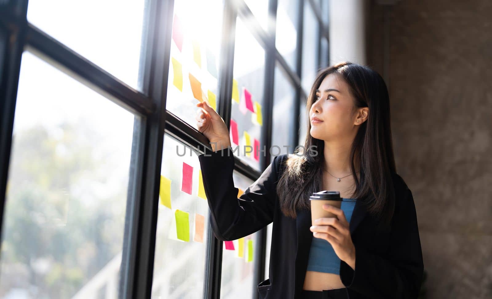 Young creative woman professional using sticky notes in glass wall to writing strategy business plan to development grow to success. Happy asian woman working at office. by wichayada