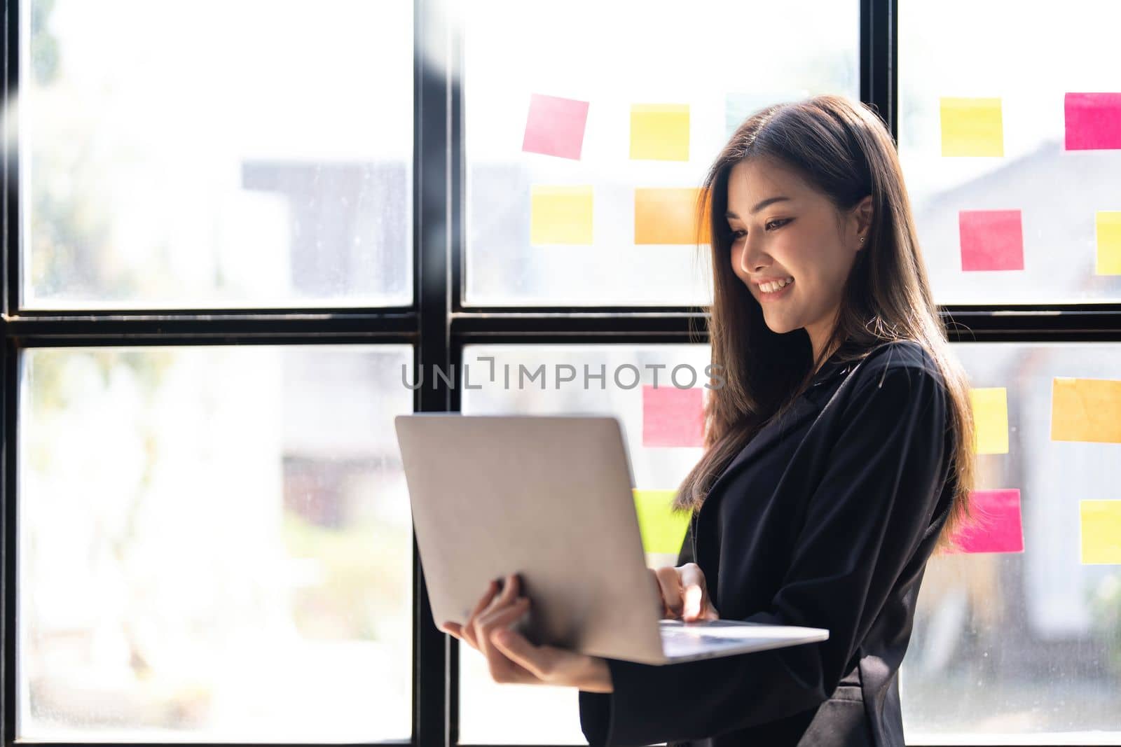 Asian freelance woman smiling paste post-it on the wall to record for work and using on laptop on wooden table at home. Entrepreneur woman working for her business at home. Business concept. by wichayada