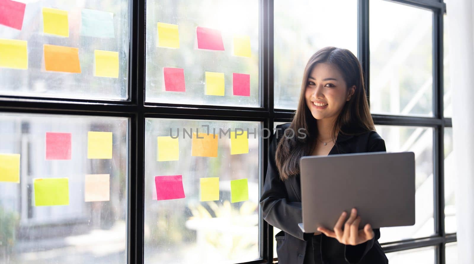 Asian freelance woman smiling paste post-it on the wall to record for work and using on laptop on wooden table at home. Entrepreneur woman working for her business at home. Business concept. by wichayada