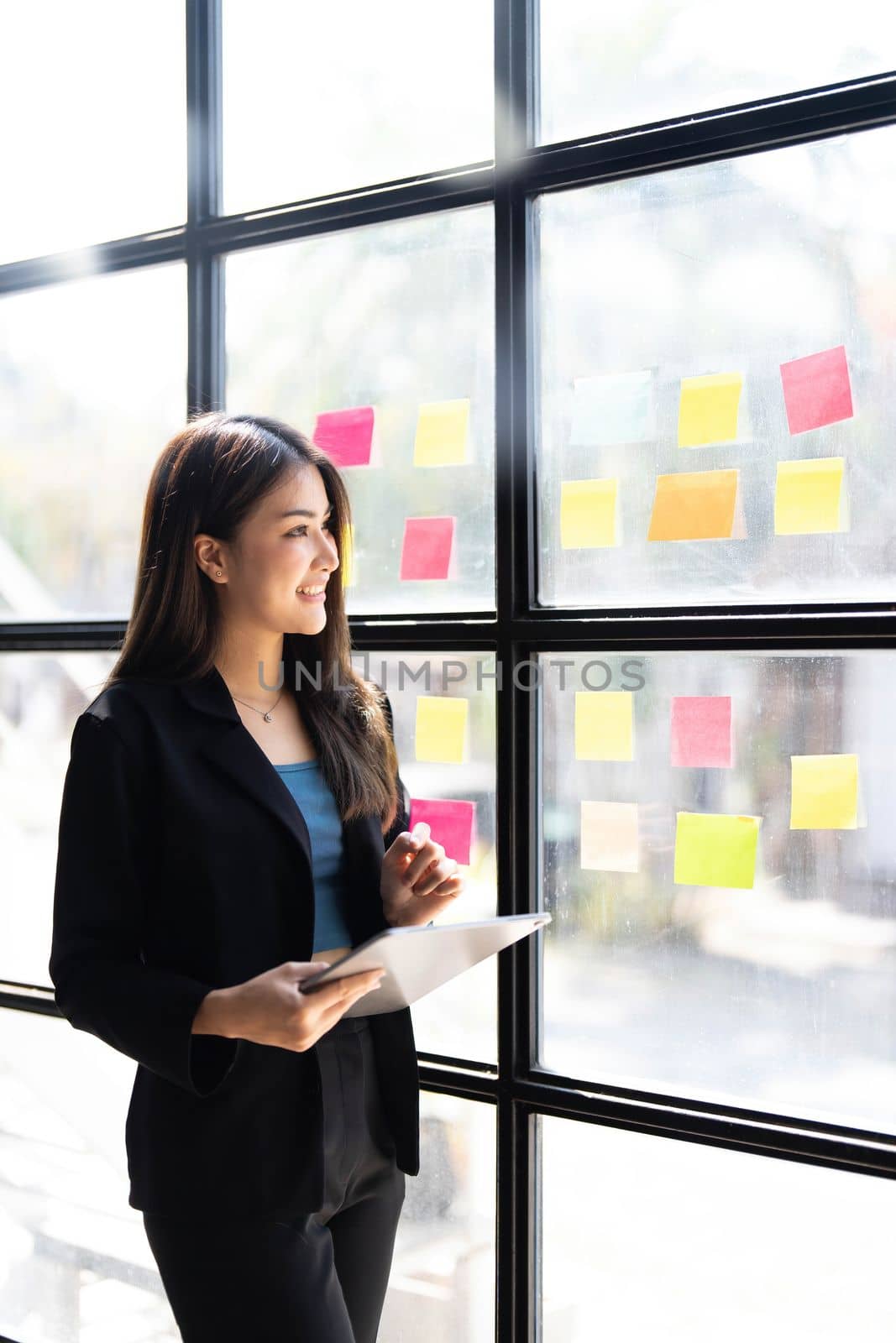Young creative woman professional using sticky notes in glass wall to writing strategy business plan to development grow to success. Happy asian woman working at office. by wichayada