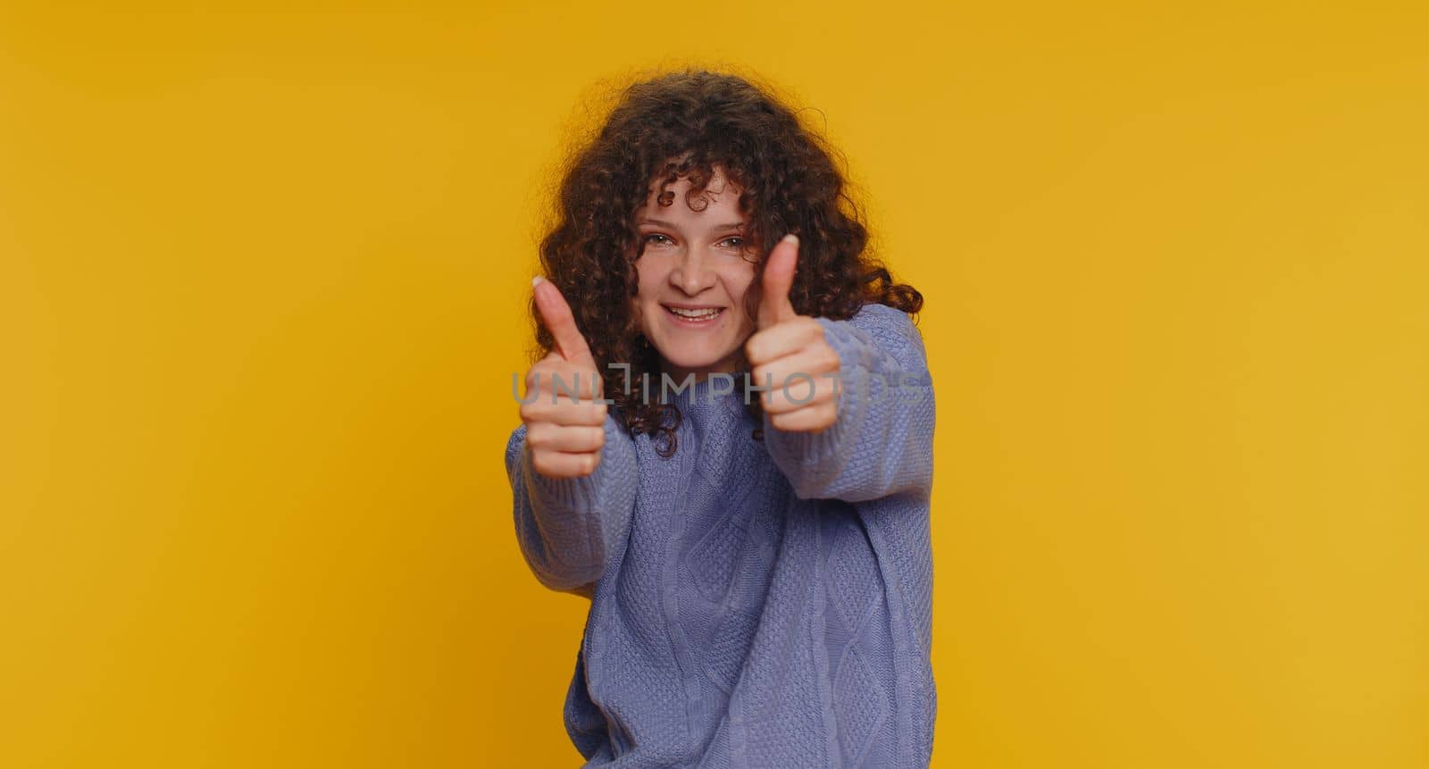 Like. Pretty curly haired woman in sweater raises thumbs up agrees with something or gives positive reply recommends advertisement likes good. Young girl isolated alone on yellow studio background