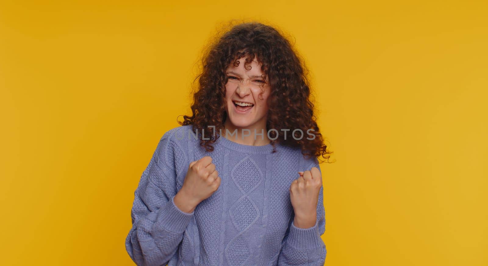 Happy joyful curly haired woman shouting, raising fists in gesture I did it, celebrating success, winning, birthday, lottery jackpot goal achievemen. Adult girl indoors on yellow studio background
