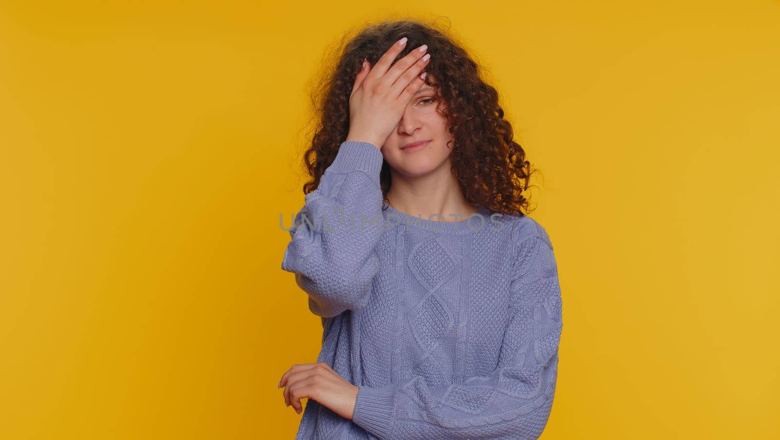 Face palm. Shame on you. Upset curly haired woman making face palm gesture, feeling bored, disappointed in result, bad news, ridiculous joke. Young teen girl isolated alone on yellow studio background