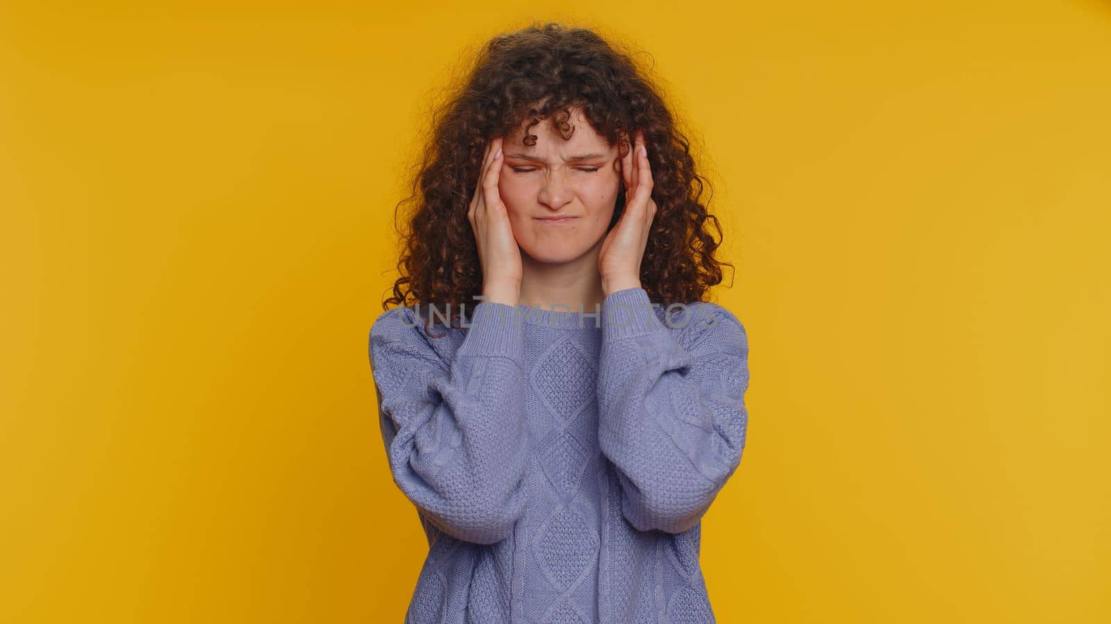 Hipster young curly haired woman in sweater rubbing temples to cure headache problem, suffering from tension, migaine, stress, grimacing in pain, high blood pressure isolated on yellow wall background