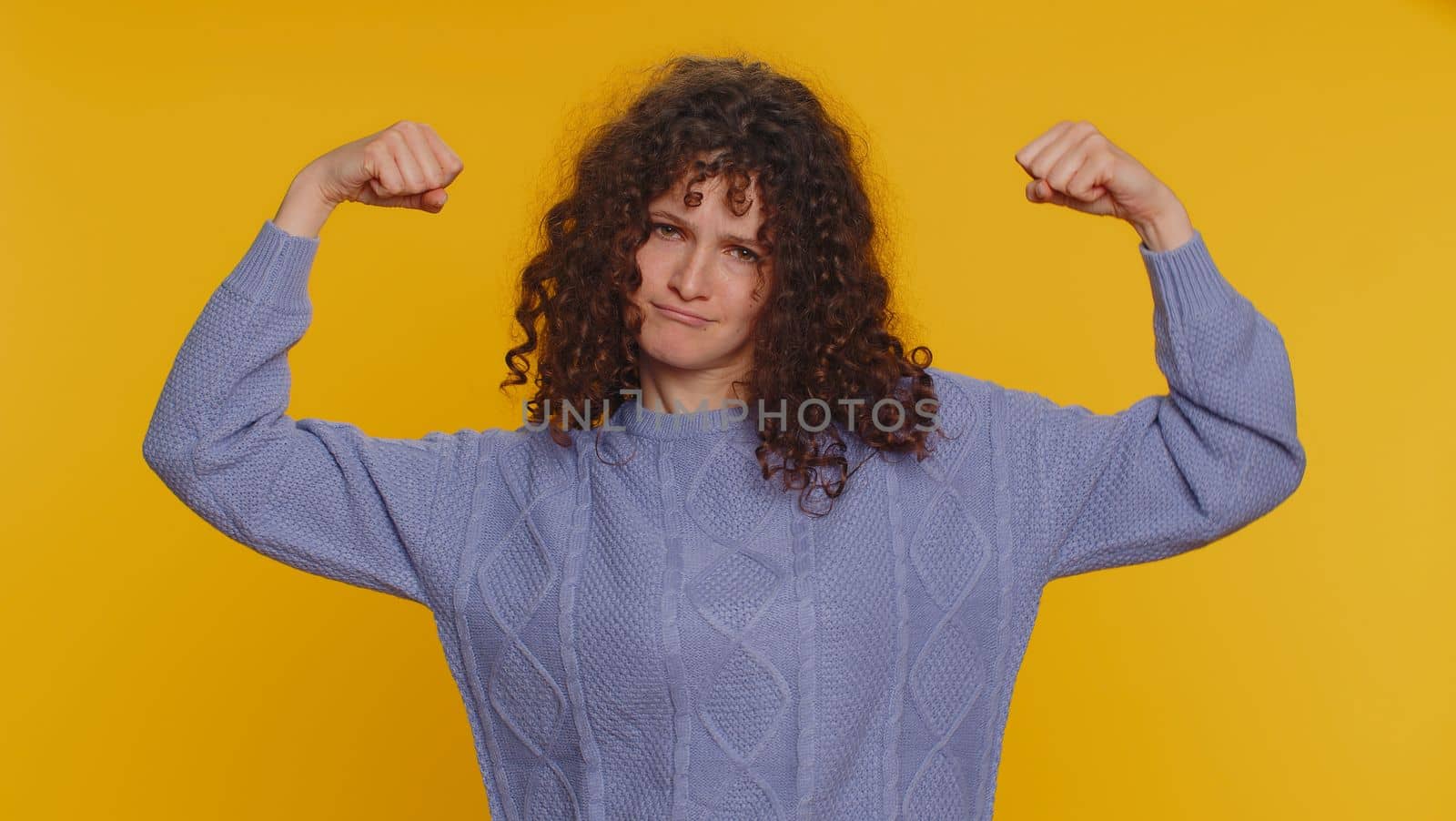 I am strong and independent. Curly haired woman showing biceps, looking confident, feeling power strength to fight for rights, energy to gain success win. Young teen girl on yellow studio background