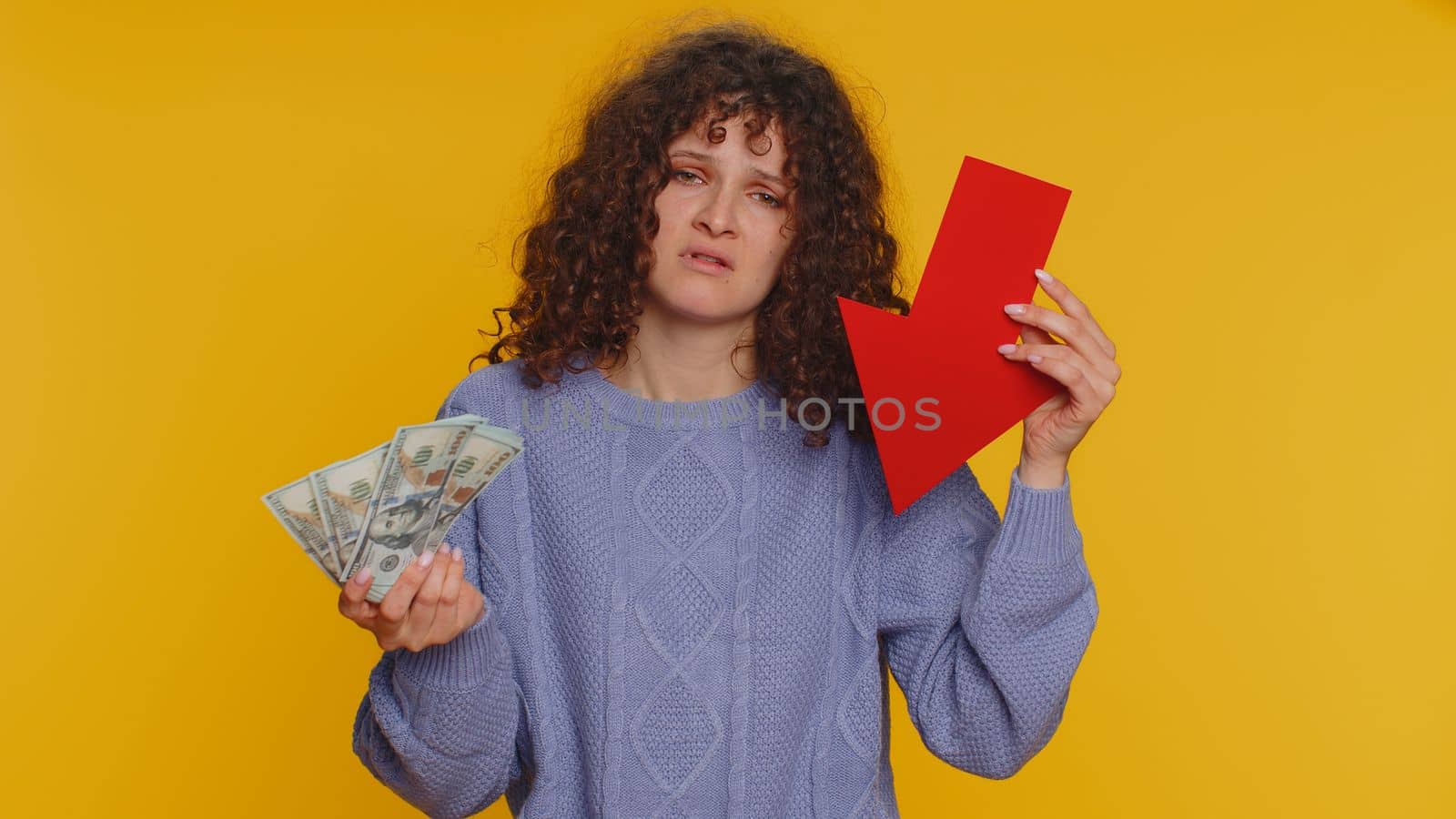 Upset curly haired woman showing red arrow pointing down, concept of downgrade, unsuccessful business, fall of stock market money dollar exchange rate bankruptcy fail. Young girl on yellow background