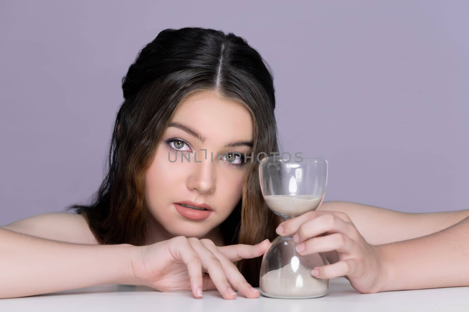 Concept of skincare and anti-aging beauty care with charming young plum girl closeup portrait holding hourglass on isolated background. Fresh and healthy face skin girl with sand glass for cosmetology
