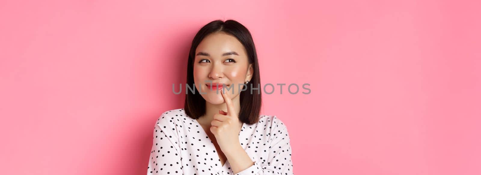 Beauty and lifestyle concept. Close-up of dreamy asian girl smiling, looking left and thinking, making choice, standing over pink background.