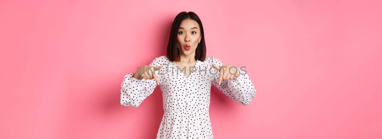 Impressed asian woman in dress pointing fingers down, look there gesture, check out discounts and sales, standing amazed over pink background.