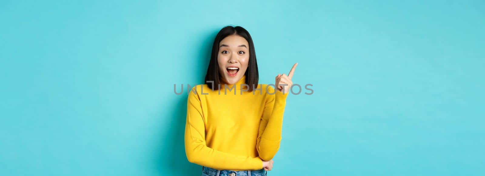 Shopping concept. Attractive korean woman smiling amazed, pointing finger left, showing good deal banner, standing against blue background.